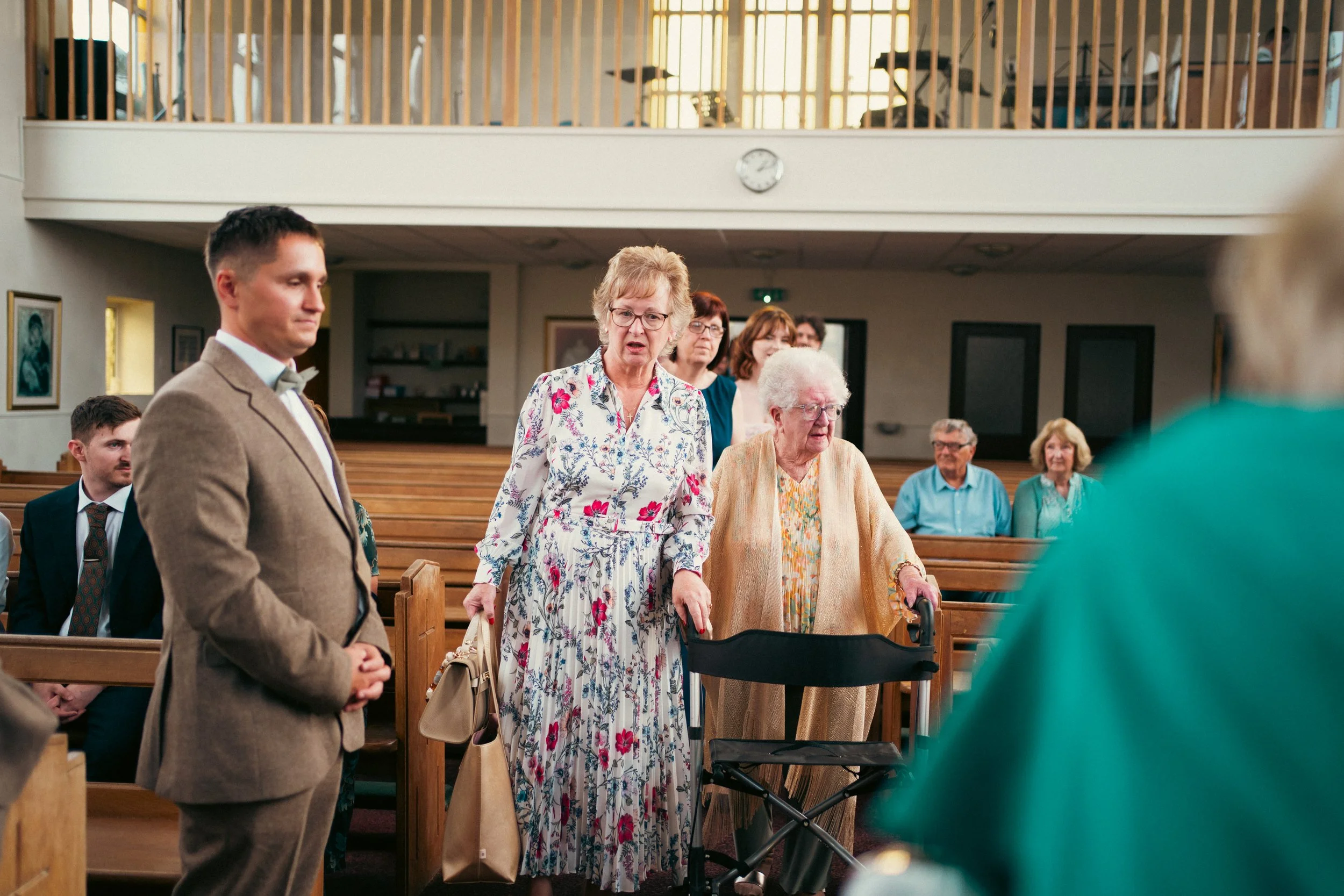 A group of elderly women and men gather inside a church during a service, with some standing and some seated, with wooden pews and religious artwork on the walls.