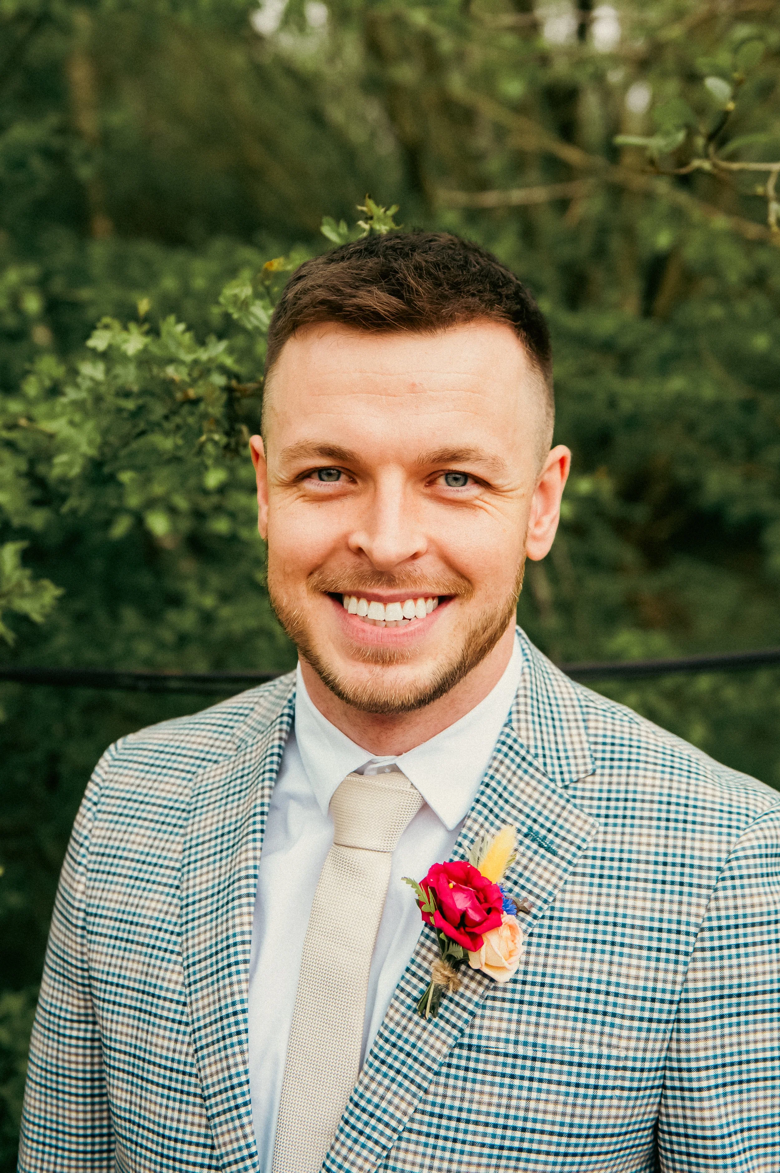 A smiling man with short dark hair and light beard, dressed in a checkered blazer, white shirt, and beige tie, standing outdoors with green trees in the background, wearing a pink and yellow boutonniere.