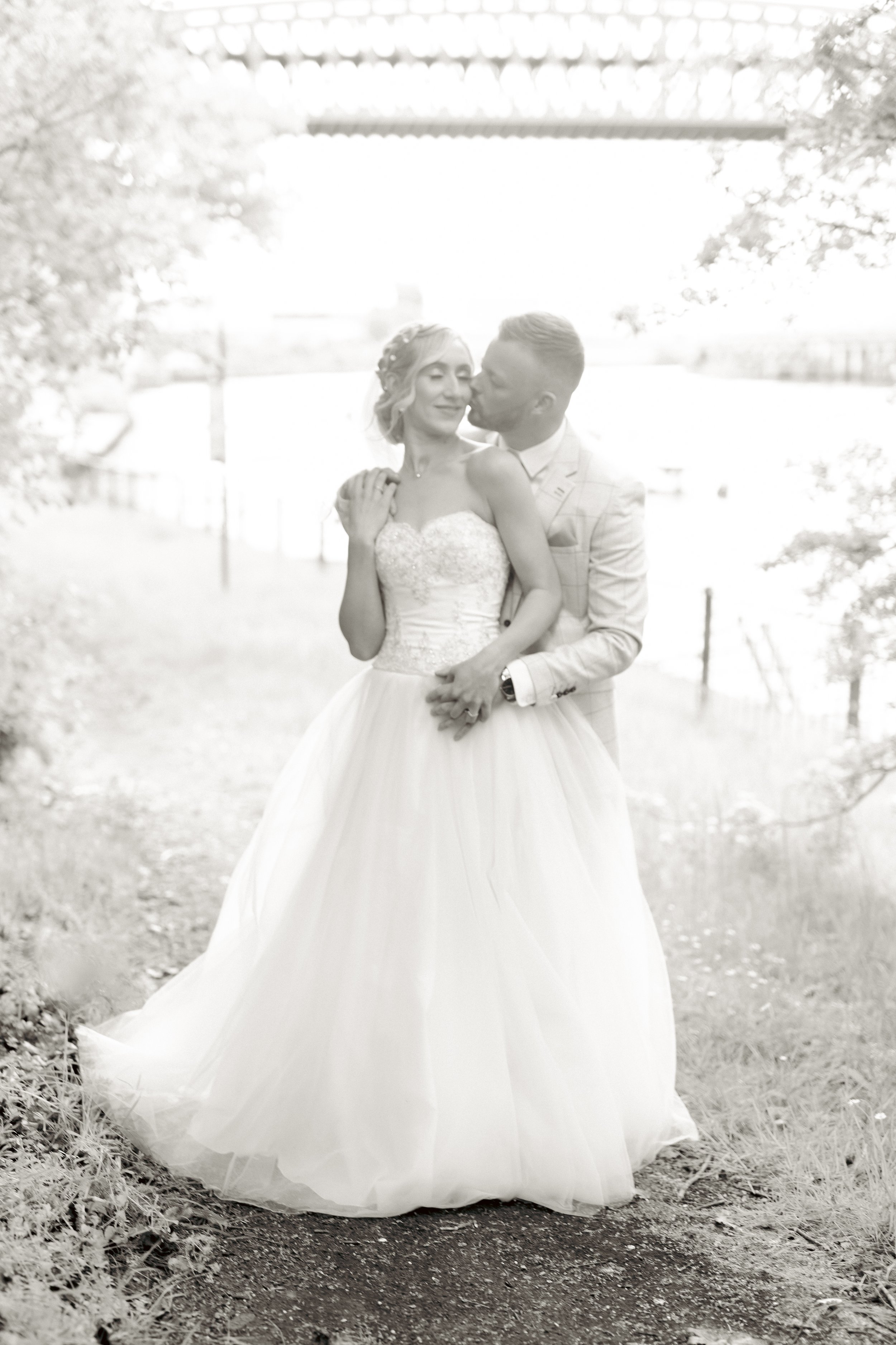 A couple in wedding attire standing outdoors, with the groom kissing the bride on her cheek, under a bridge, surrounded by trees.