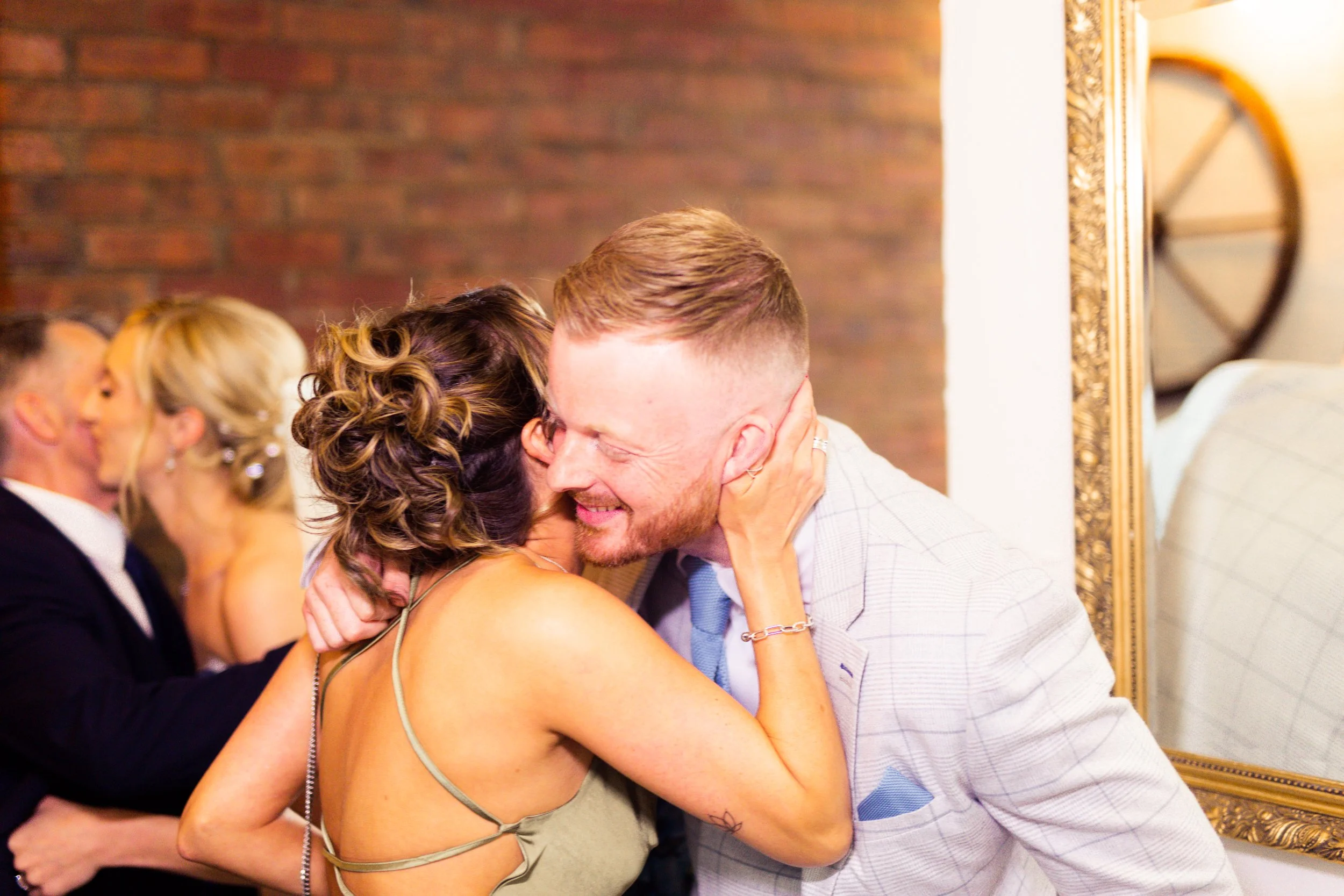 Two couples kissing at a celebration, with a woman hugging and kissing a man dressed in a light suit, and another couple kissing in the background.