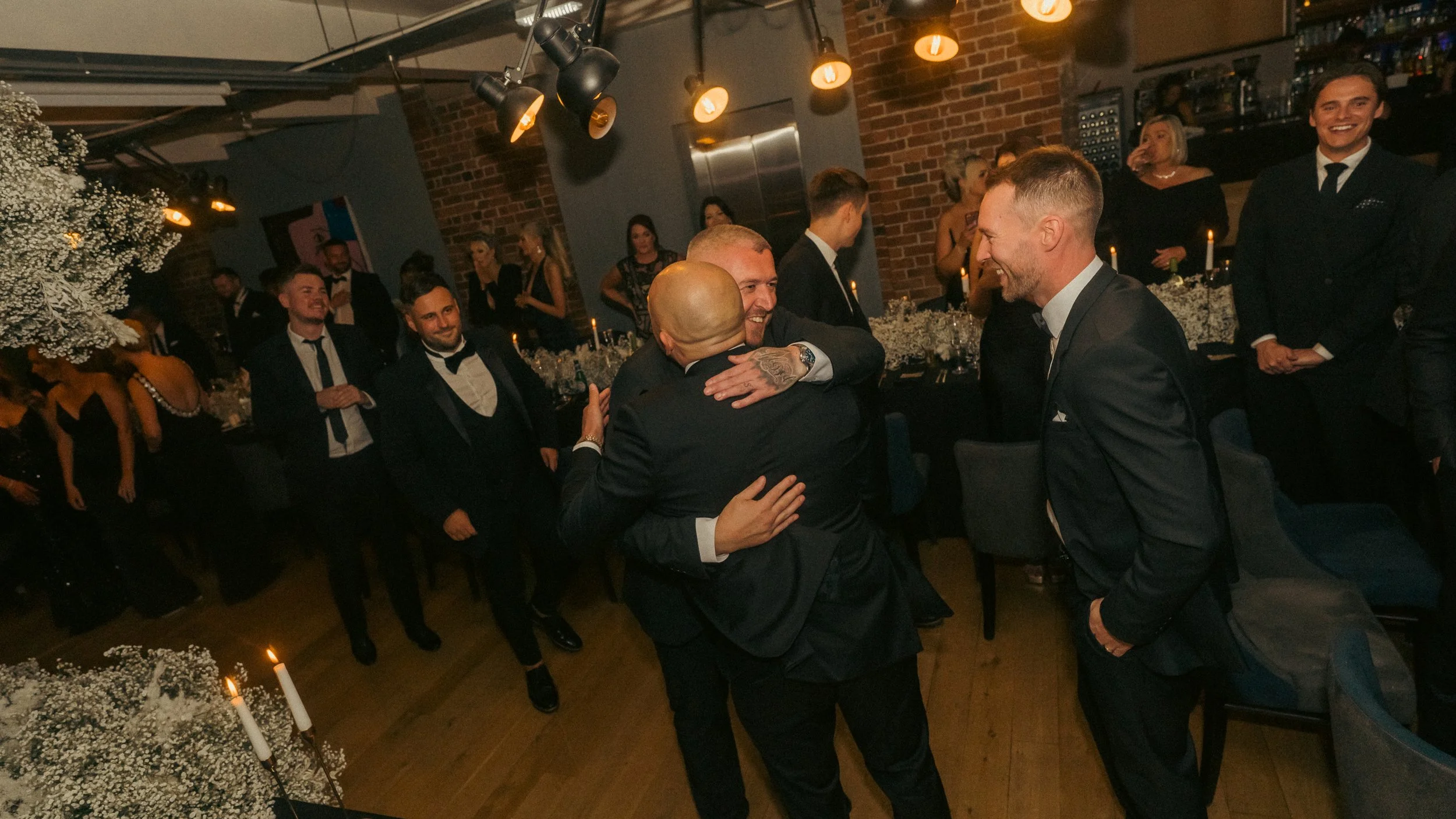 A group of men in formal suits at a celebration, with two men hugging in the center, surrounded by other smiling men and women in elegant attire, decorated with candles, flowers, and exposed brick walls.
