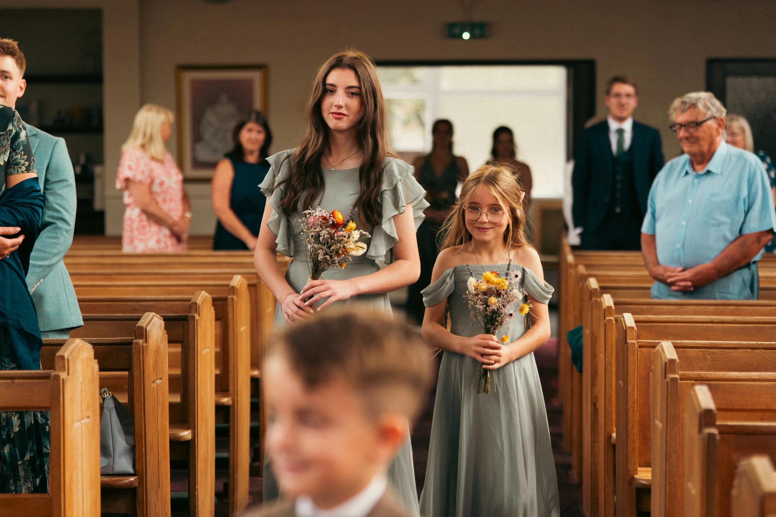 Two young girls, one taller and older, the other shorter and younger, walking down the aisle of a church during a wedding ceremony, each holding a bouquet of flowers. Other guests are standing and watching in the background.