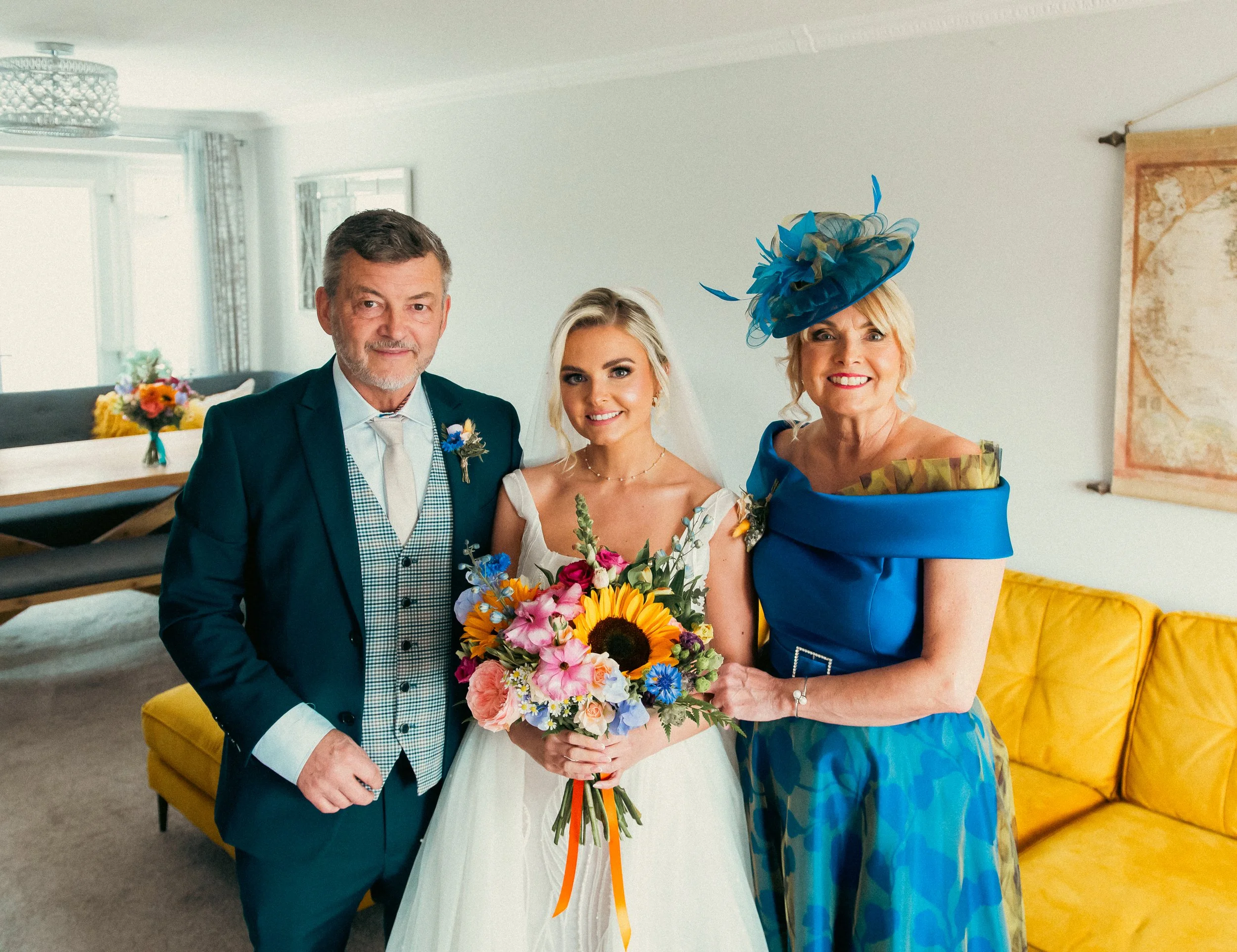 A bride in a wedding dress holding a colorful bouquet standing with an older man in a suit and a woman in a blue dress with a large matching hat inside a living room.