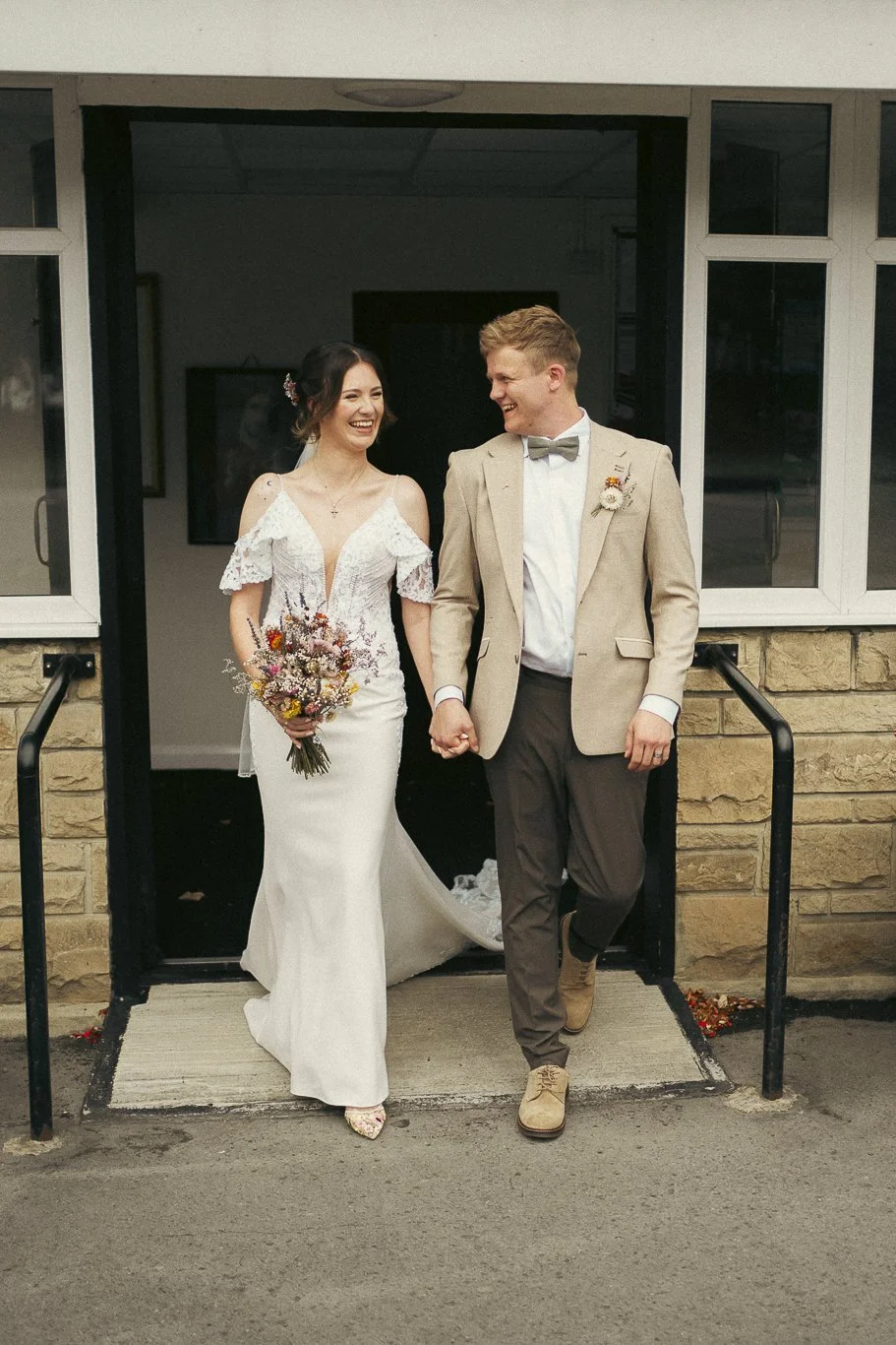 A newlywed couple holding hands and smiling as they walk out of a building. The bride is wearing a white lace wedding dress and holding a bouquet, and the groom is dressed in a beige suit with a bow tie. They appear happy and joyful.