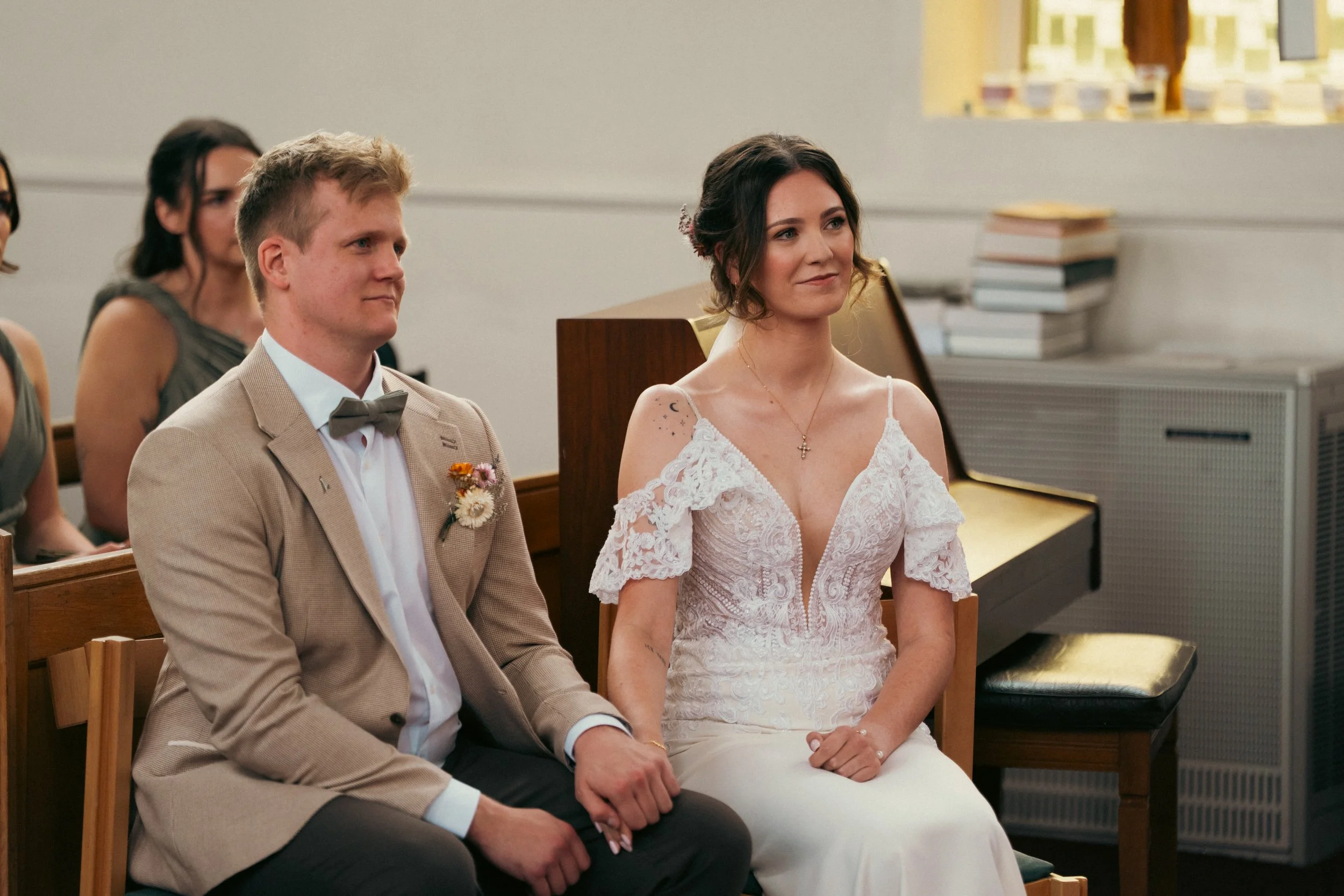 A bride and groom sitting at their wedding ceremony, holding hands, with female guests in the background