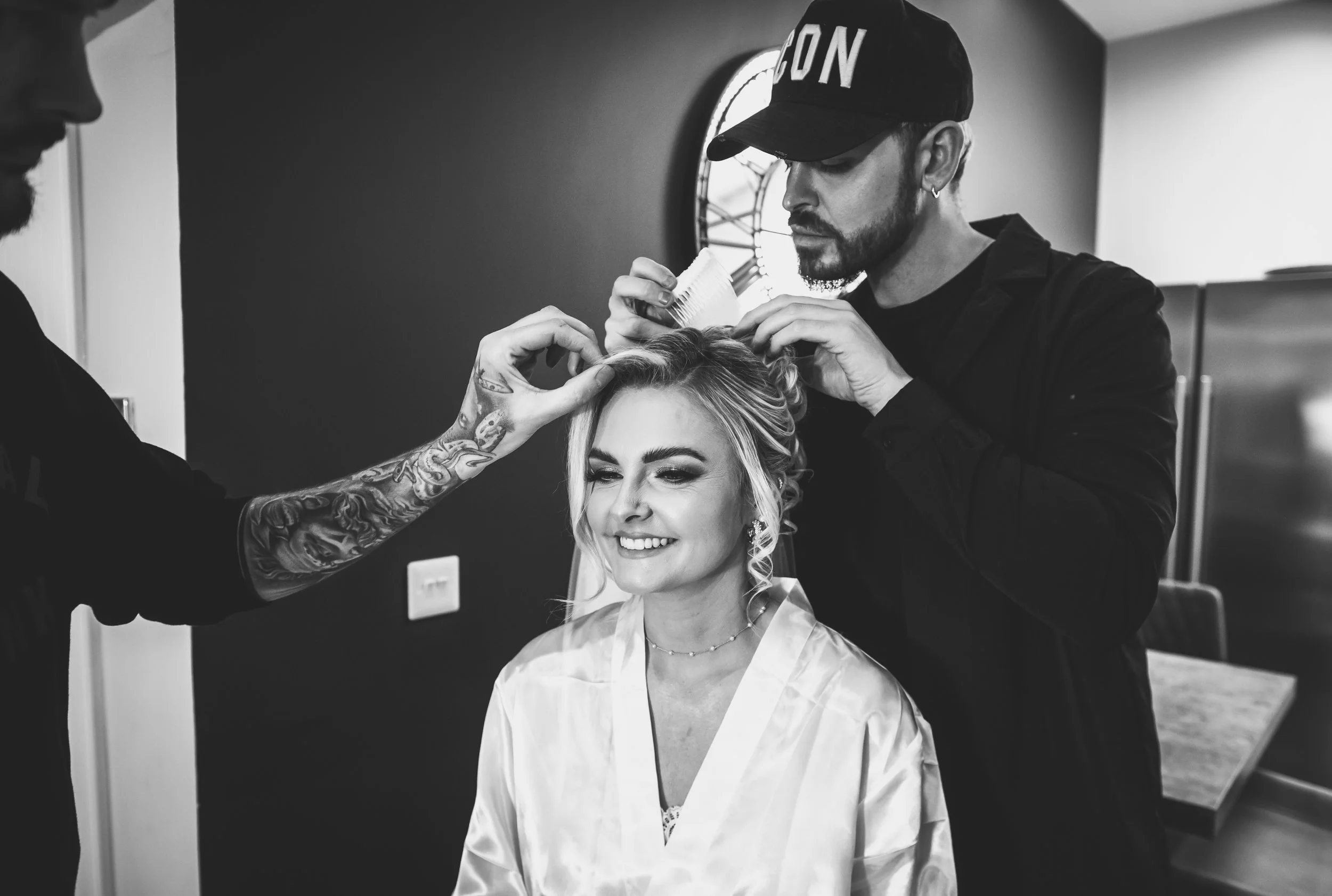 A woman getting her hair styled by a hairstylist in a salon, smiling with eyes closed, with another stylist working on her hair. The scene is in black and white.