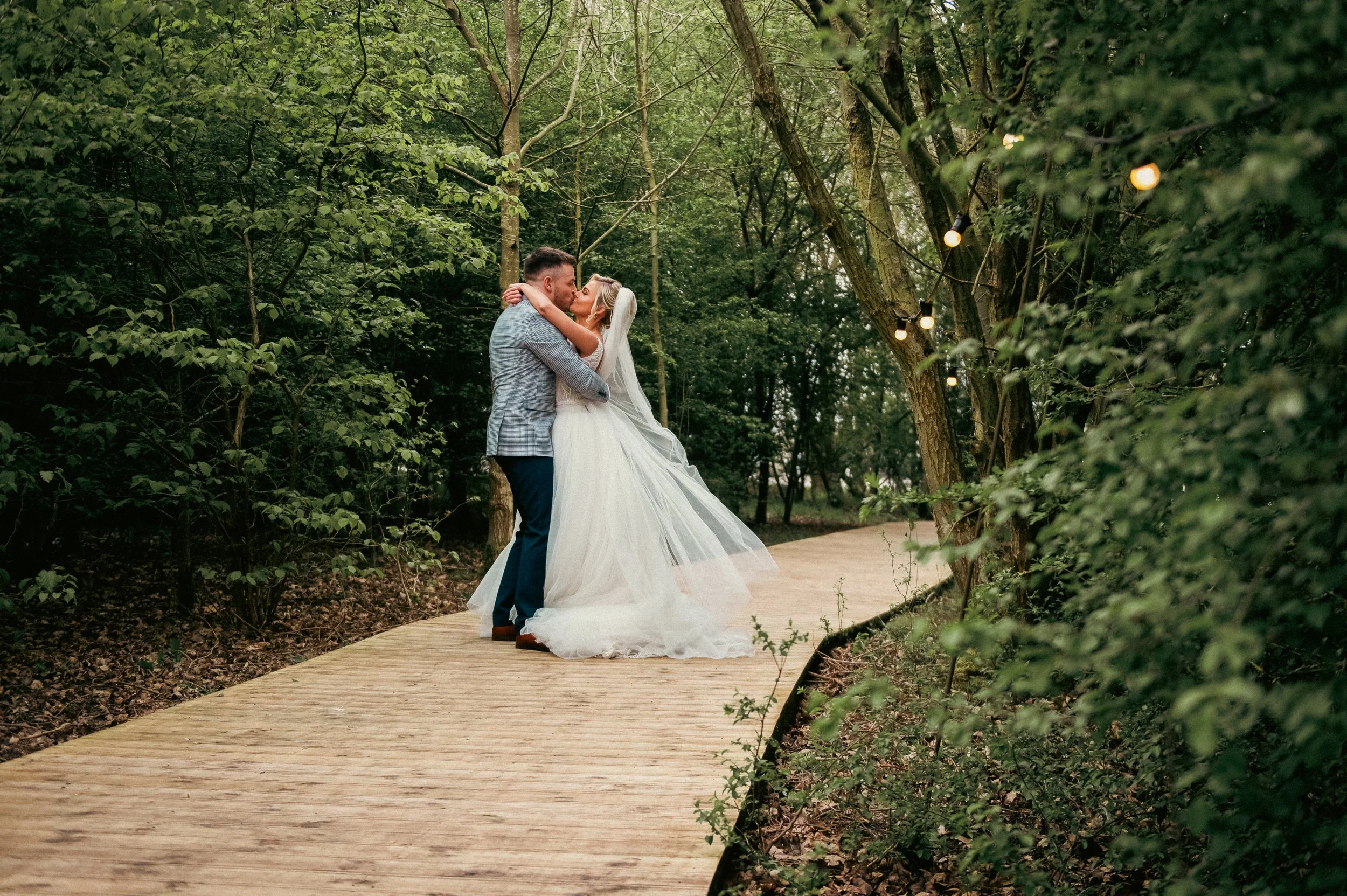 A newlywed couple sharing a kiss on a wooden pathway in a lush, green forest, with string lights hanging from the trees.