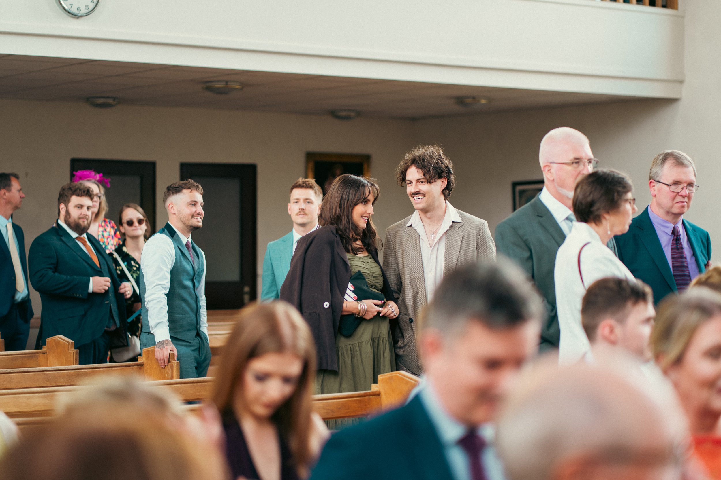 People dressed in formal attire inside a church, standing and engaging in conversations, with wooden pews and a wall clock visible.