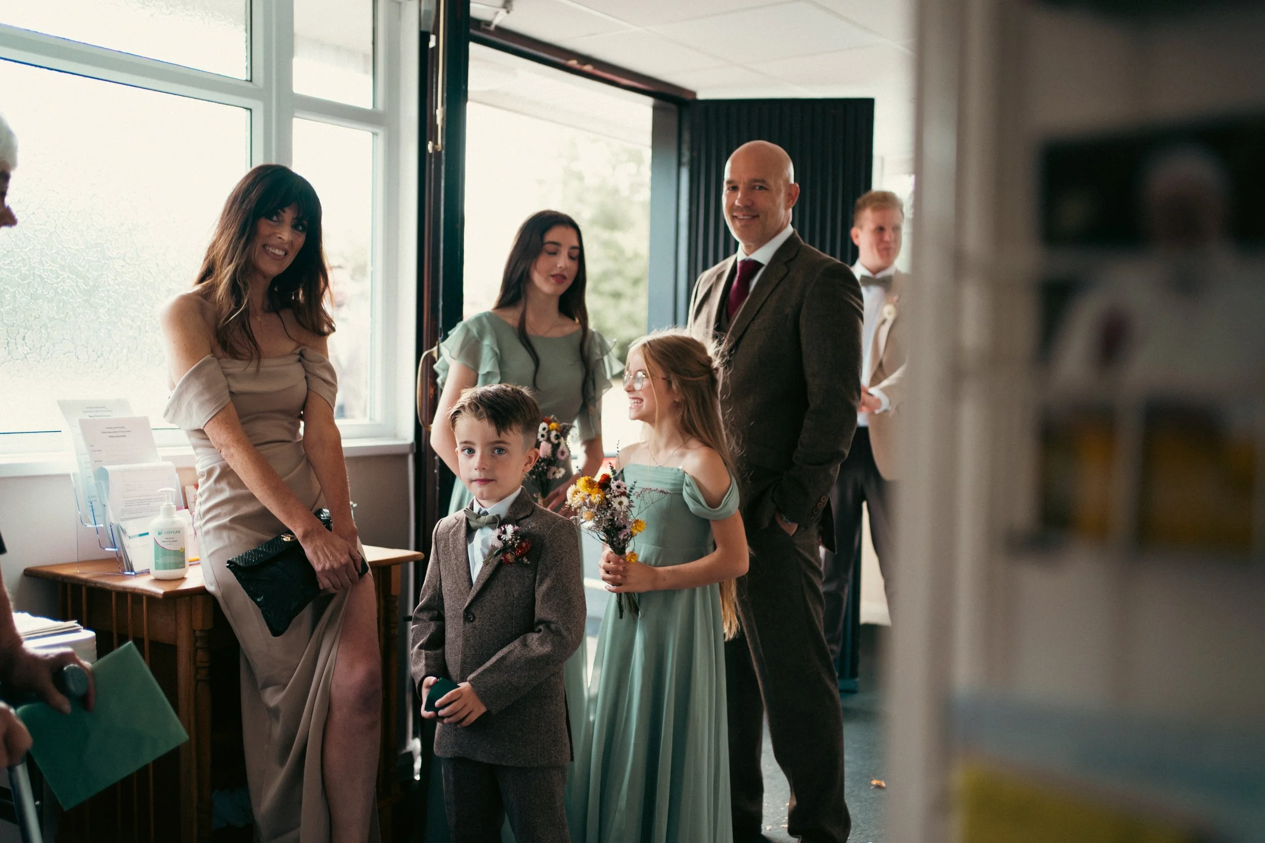 A group of people at a formal event, including children and adults, standing inside a room near a window with natural light. The children are dressed in suits and dresses, holding flowers, while the adults are dressed in formal attire.