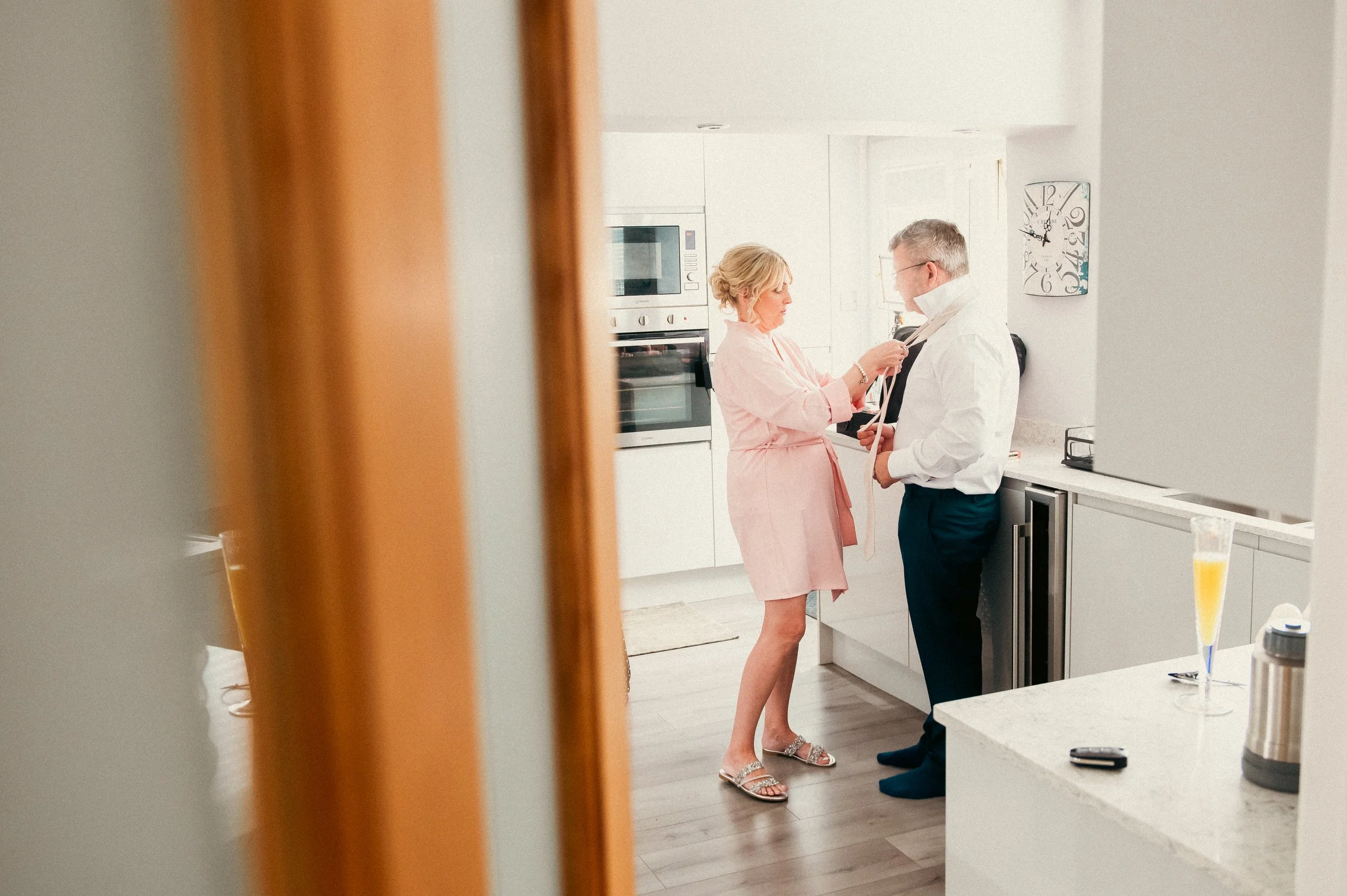 A woman in a pink dress helping a man in formal attire in a modern, white kitchen, seen through a doorway