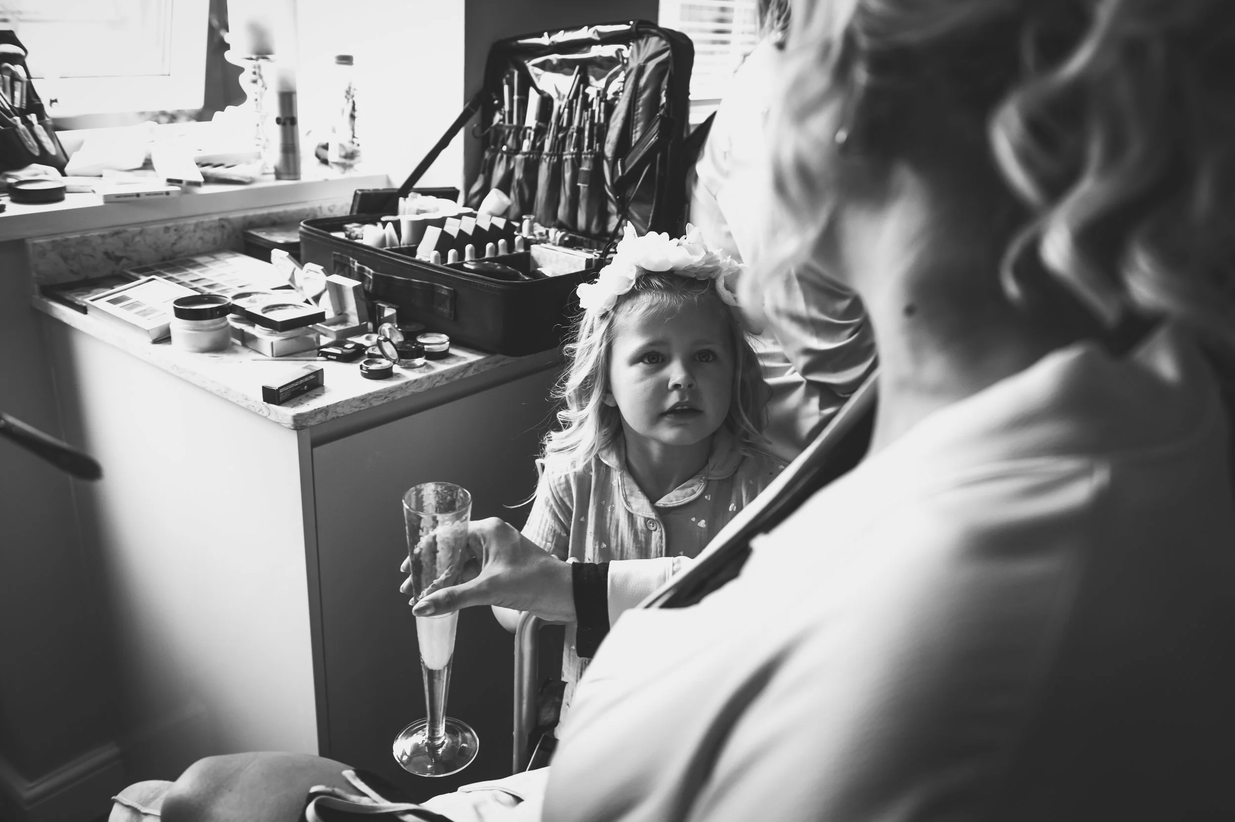A young girl with a flower headband sitting in a medical setting, looking at a person holding a champagne flute. A table with makeup and medical supplies is in the background.