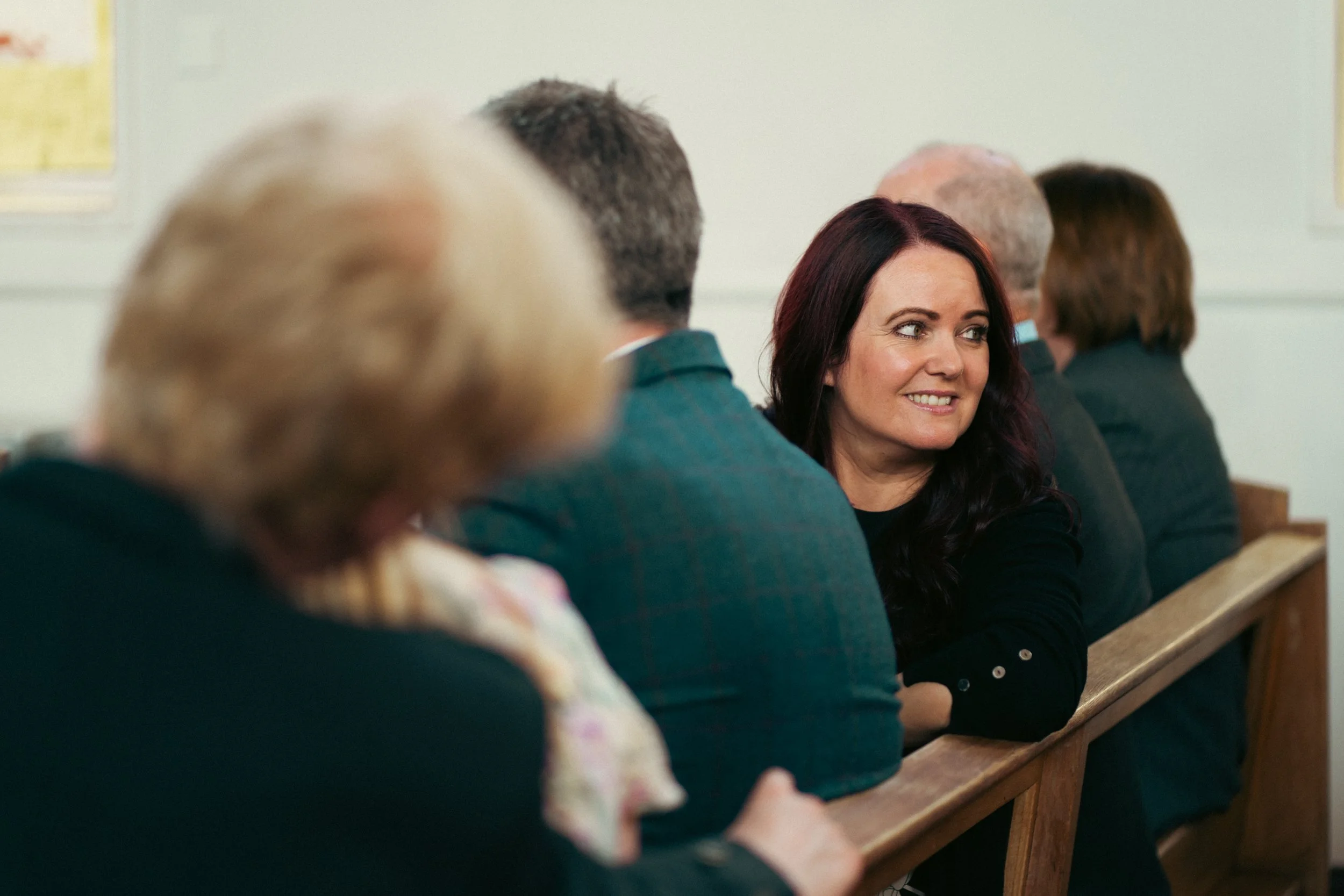 A woman with dark hair sitting in a pew at church, looking to her right and smiling.