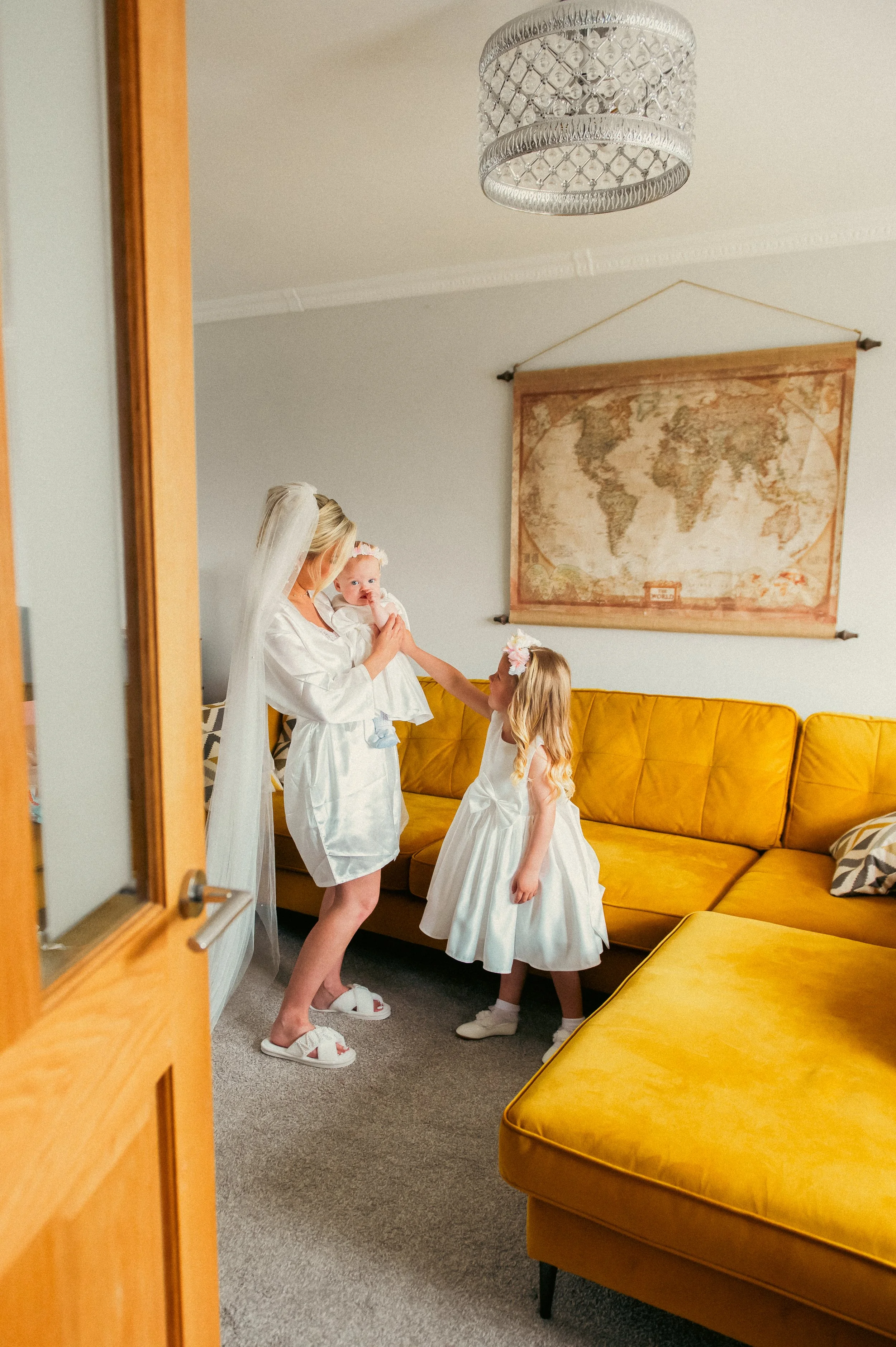 A woman, wearing a white robe and veil, holding a baby dressed in white, is being approached by a young girl in a white dress with a floral headband, inside a living room with a yellow couch and a world map on the wall.