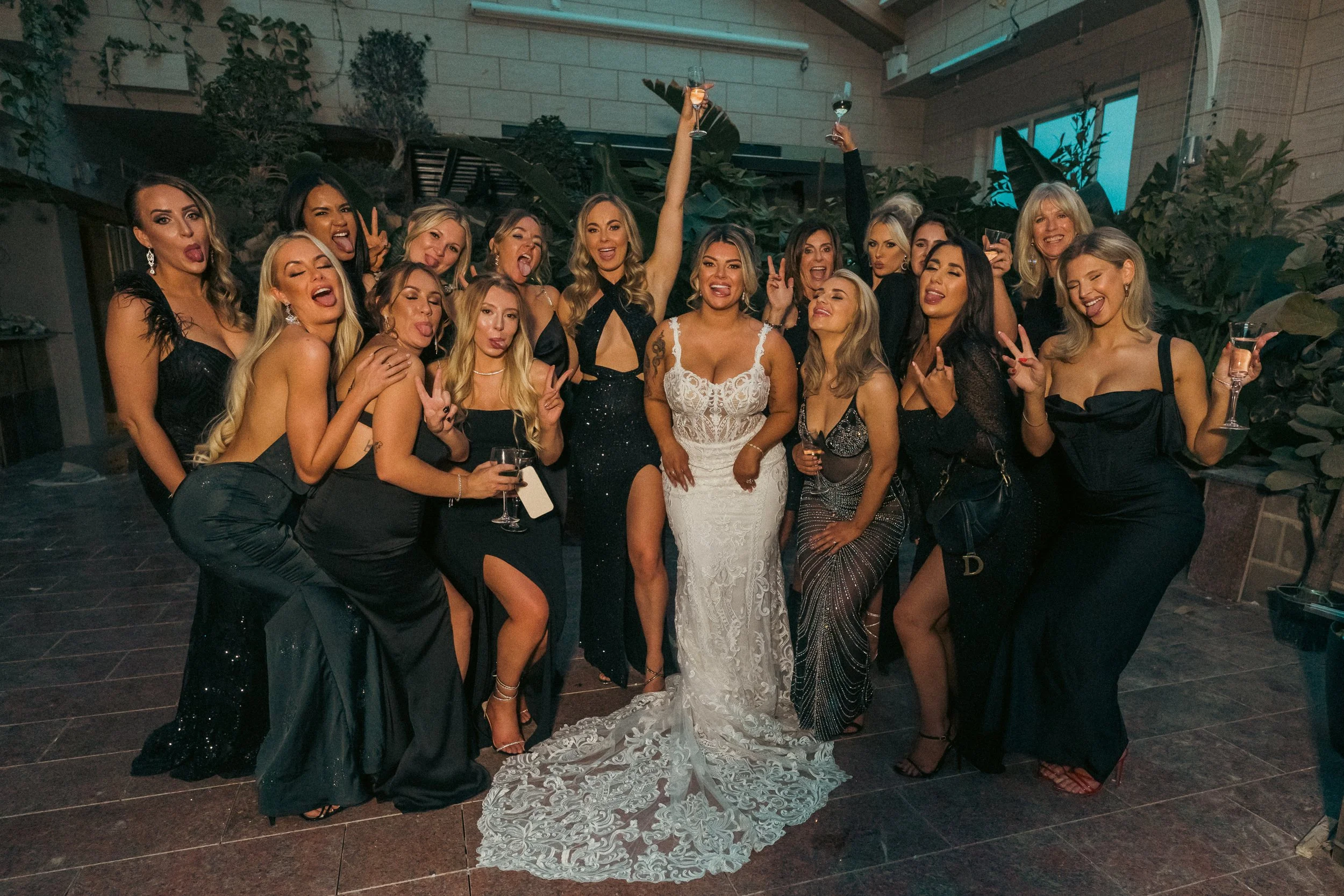 A group of women dressed in elegant evening gowns celebrating at a party, some holding glasses of wine, in a dimly lit indoor venue with plants and a high ceiling.