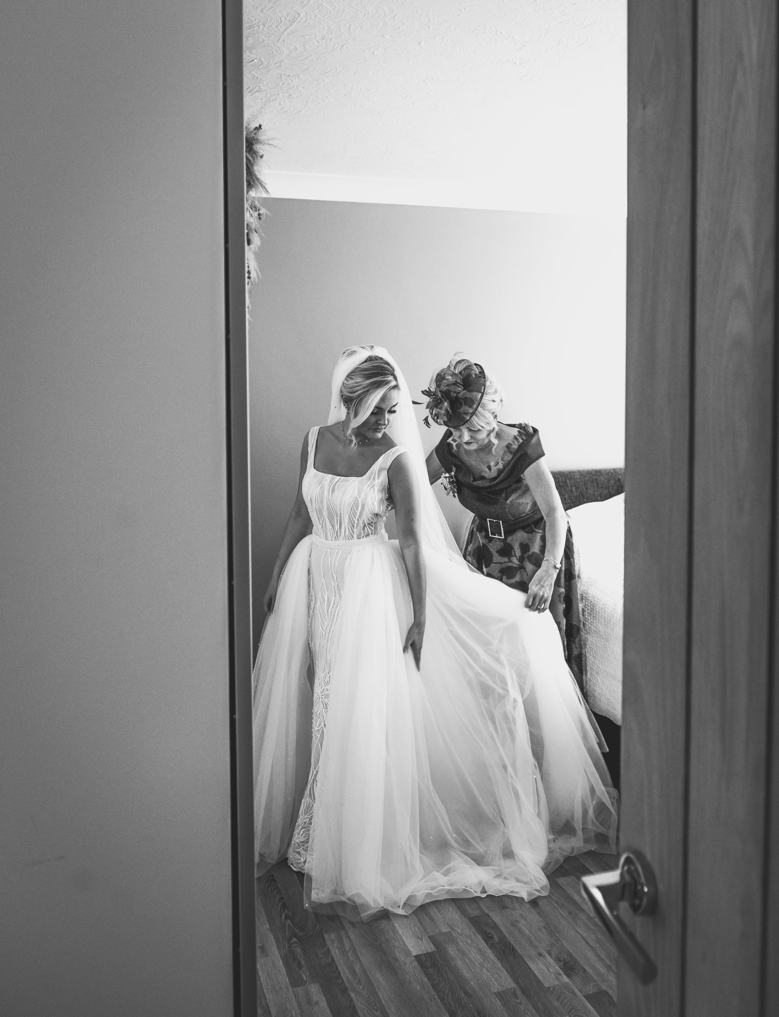 A bride in a wedding dress being assisted by a woman, possibly her mother, in a room reflected in a mirror.