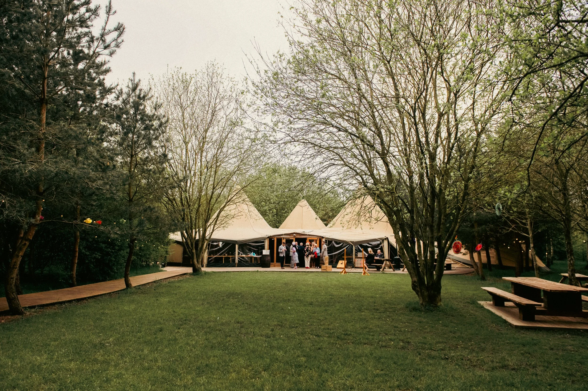 People gathering in front of a large beige tipi tent surrounded by trees and outdoor seating.