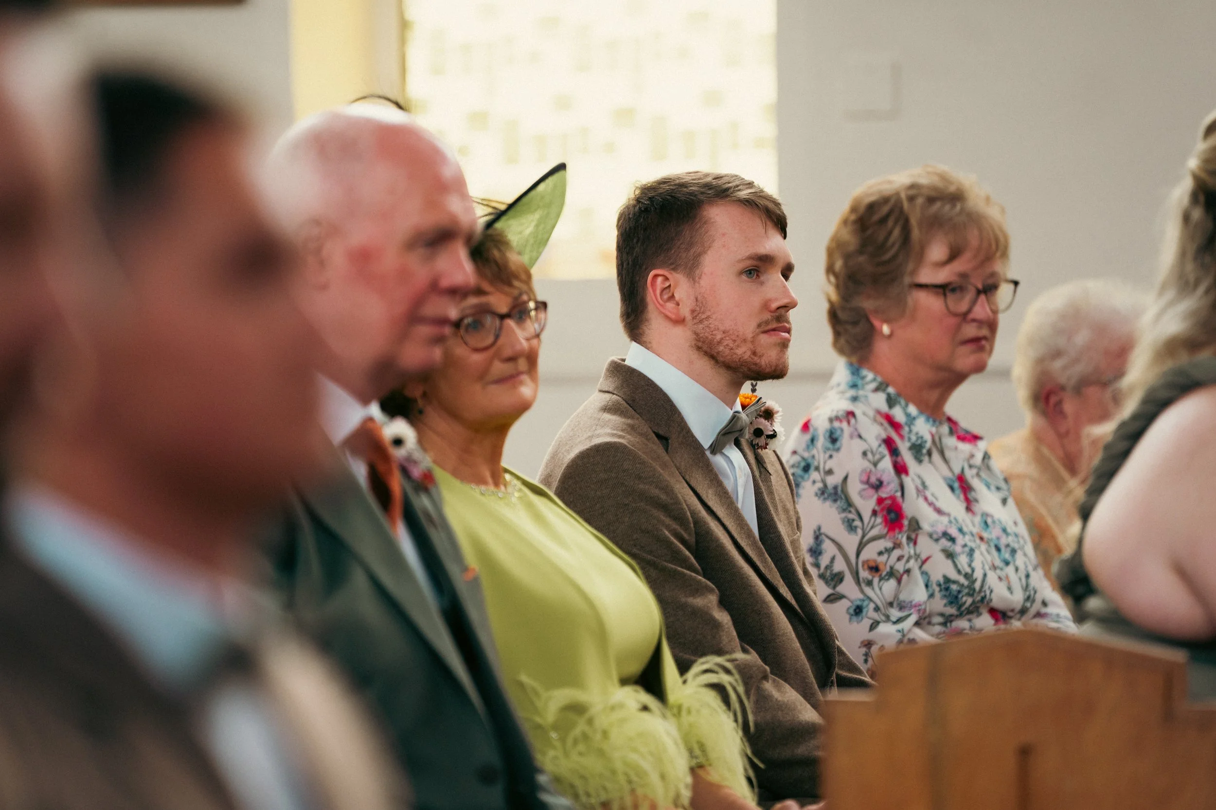 A group of people sitting in a church, attending a wedding ceremony. The group includes an older man, a woman with glasses, a young man in a brown suit, a woman with curly hair and glasses, and an elderly woman. They are seated in front of a wooden p