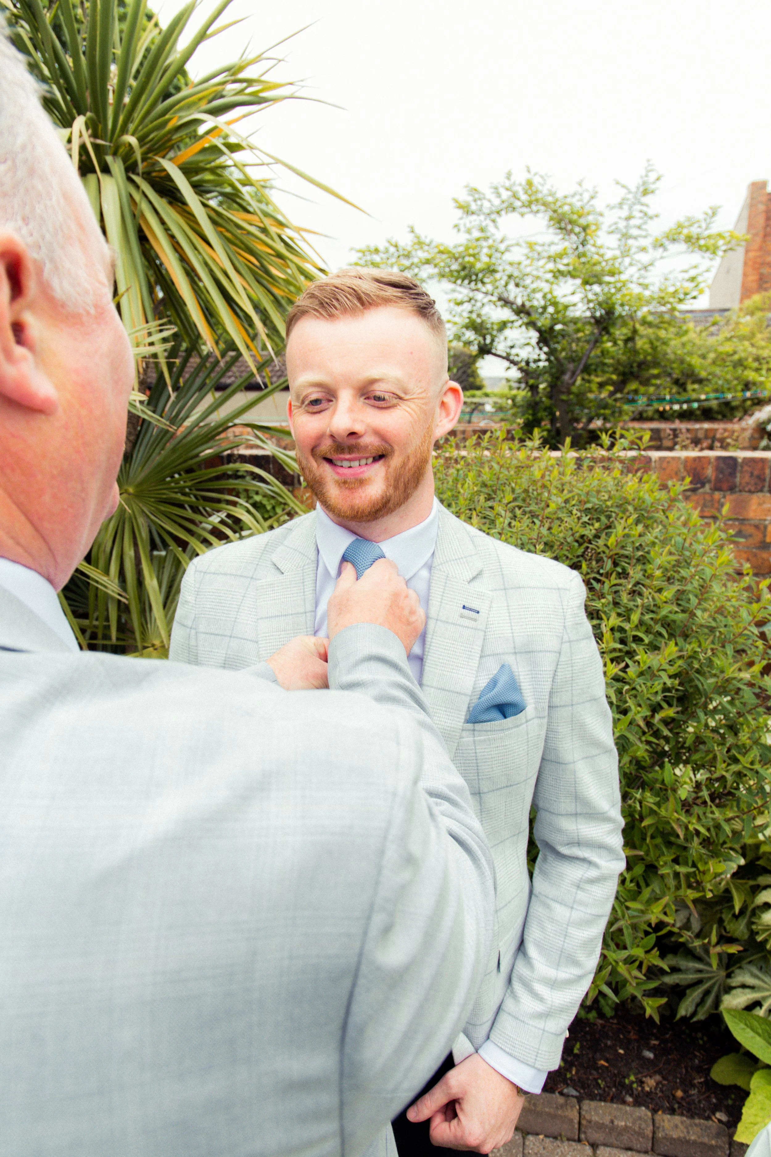 A man in a light gray plaid suit with a blue pocket square is smiling as another man adjusts his light blue tie outdoors in a backyard garden with green shrubs and trees.