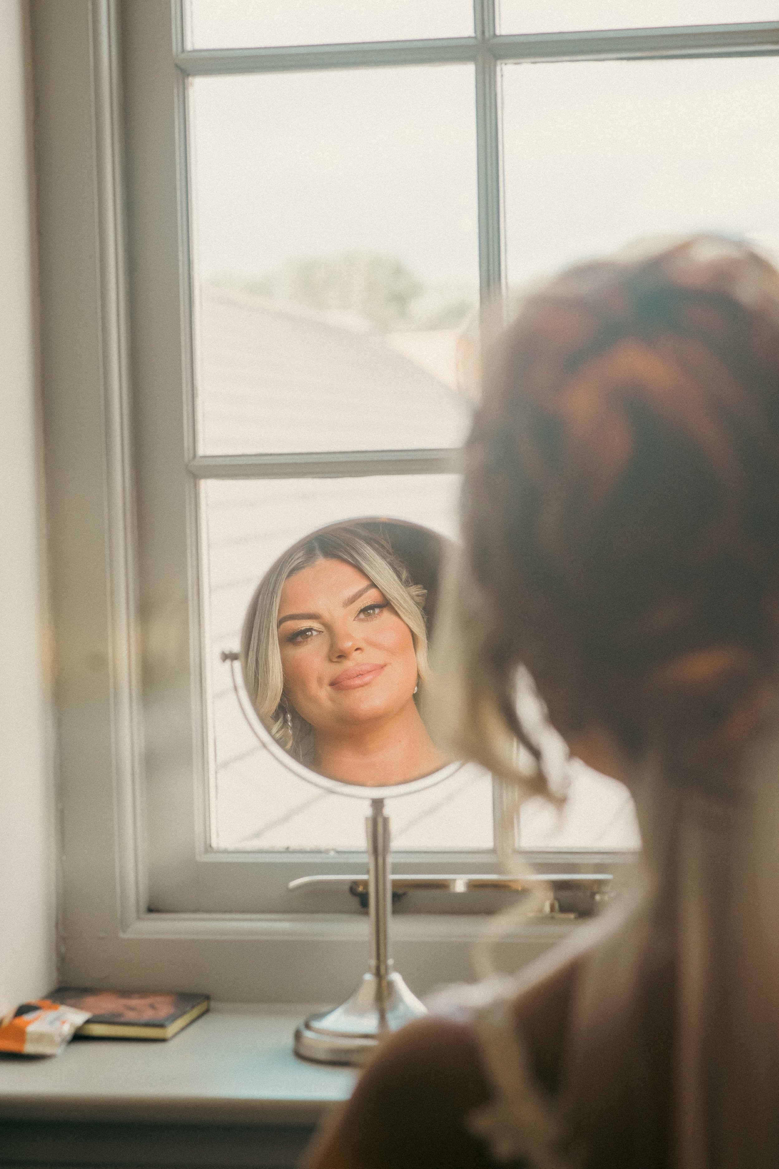 A woman with styled hair sitting in front of a mirror, looking at her reflection through a small round mirror on a window sill.