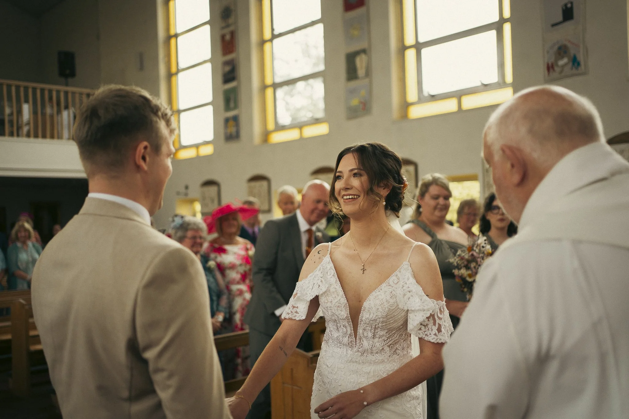 A bride and groom holding hands during a wedding ceremony inside a church, with guests smiling in the background.