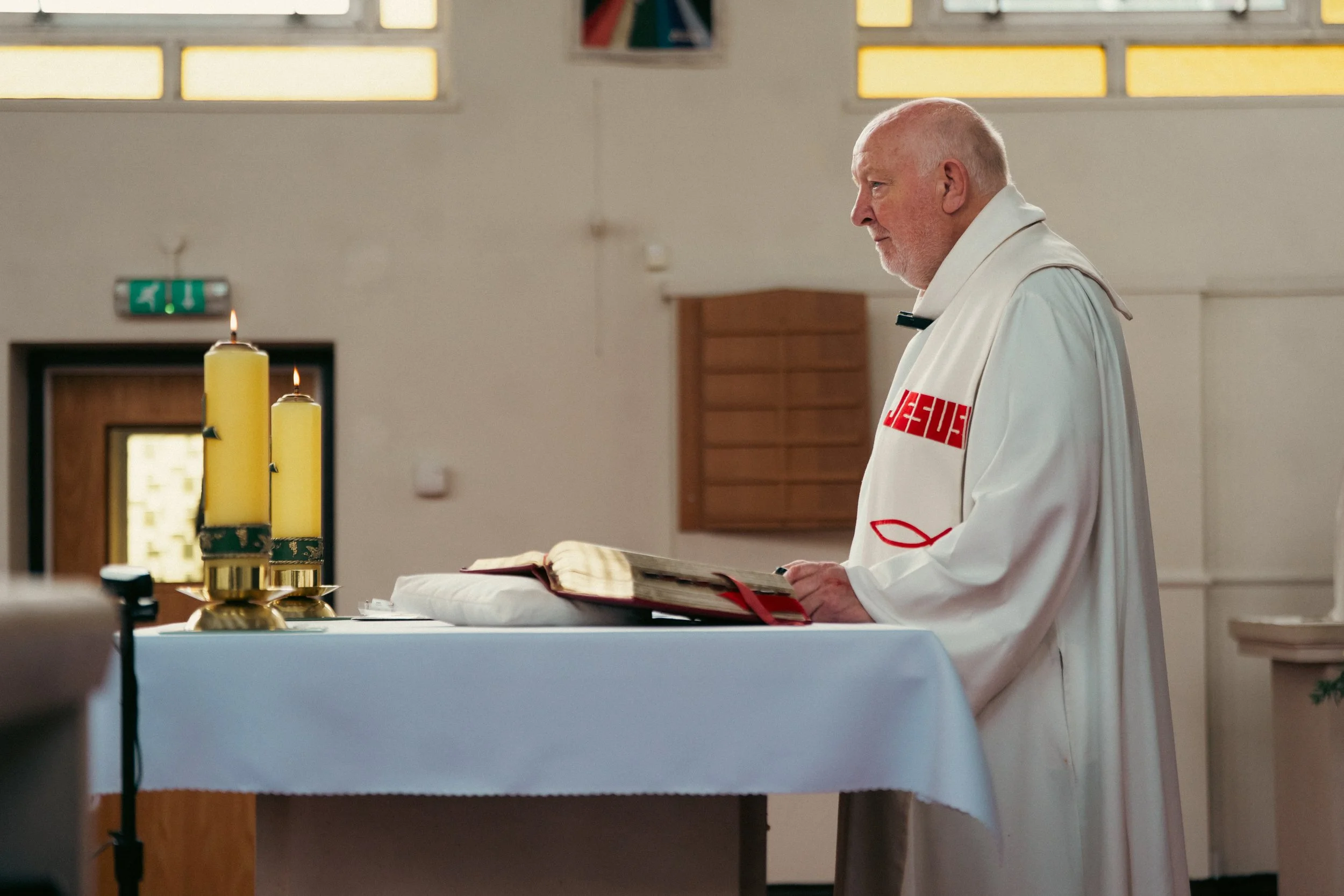 An elderly priest standing at the altar in a church, reading from a large open book, with lit yellow candles in front of him.