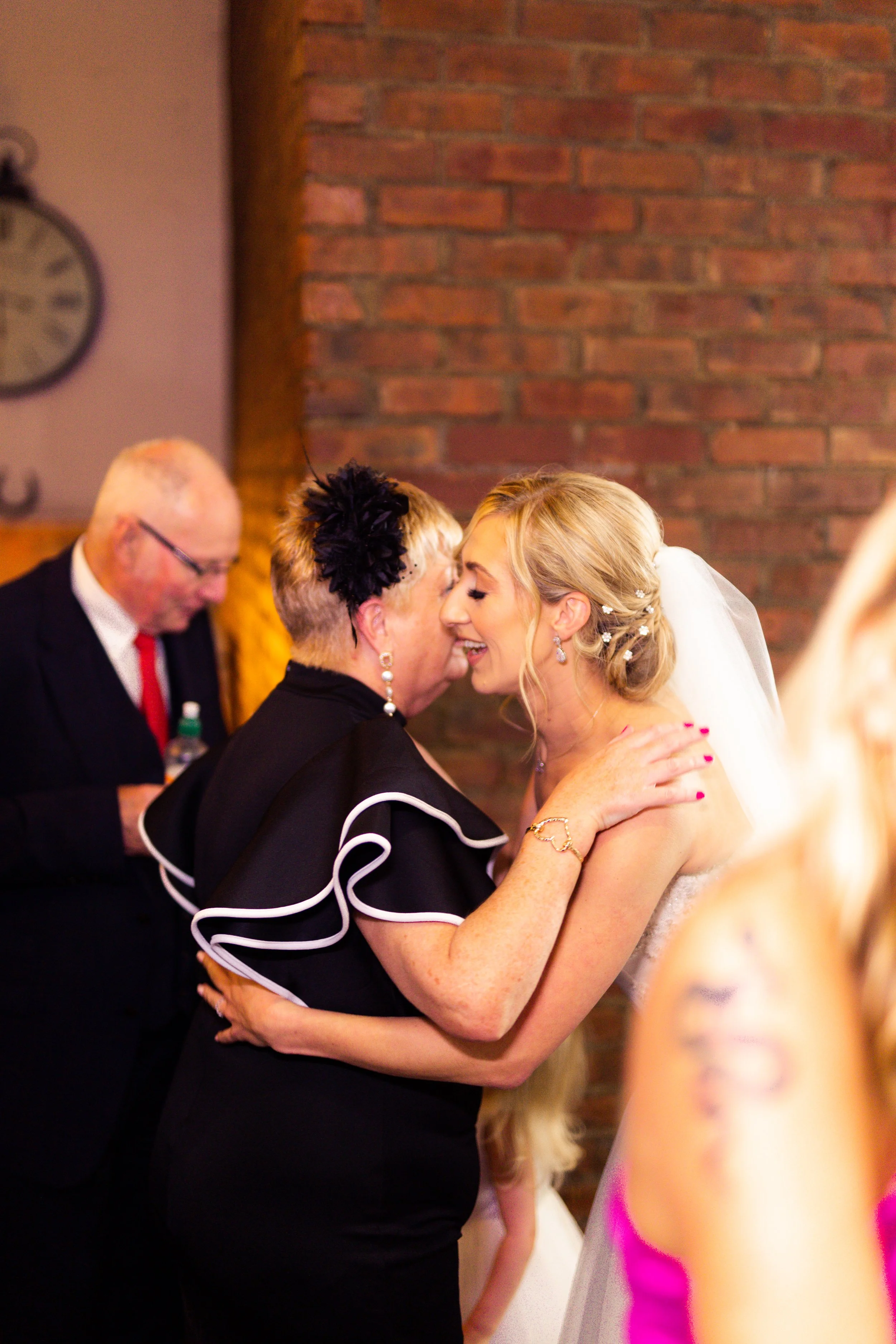 A bride and an older woman share a joyful, intimate moment, smiling and touching foreheads at a wedding reception, with a brick wall background.
