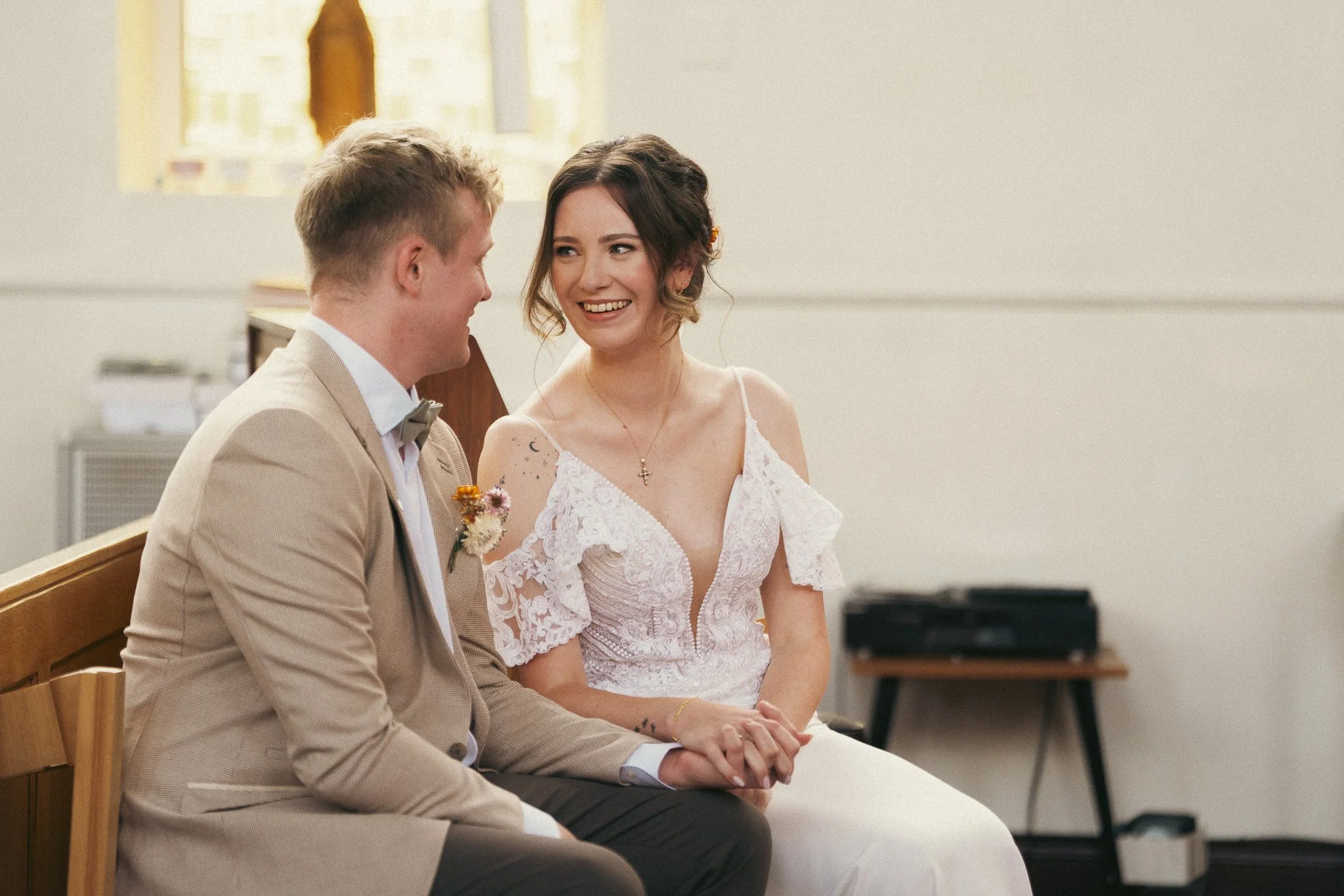 A bride and groom sit on a bench during a wedding ceremony, holding hands and smiling at each other inside a simple church with light-colored walls.