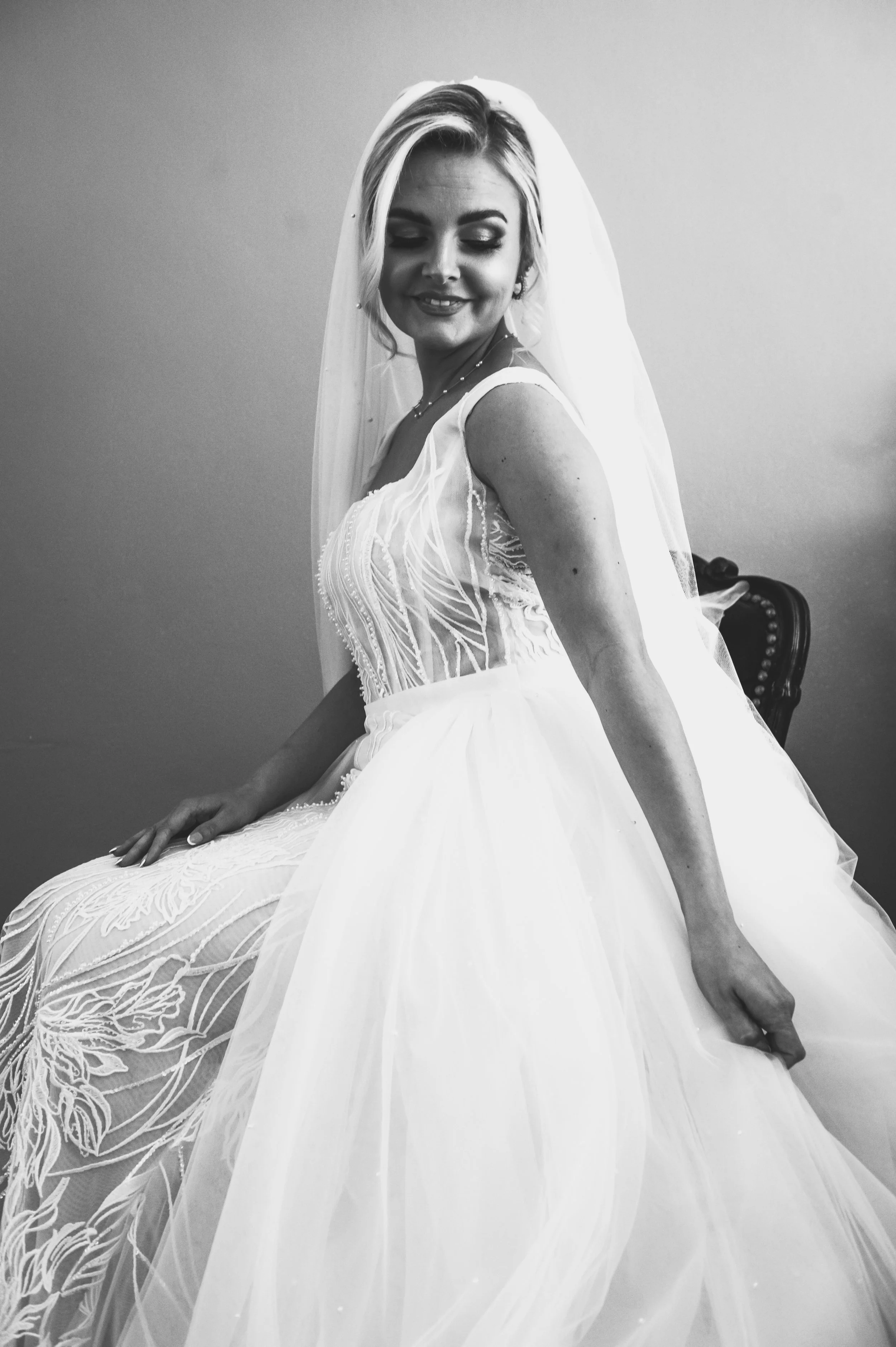 Black and white photo of a smiling woman in a wedding dress with a veil, sitting on a chair with a plain wall background.