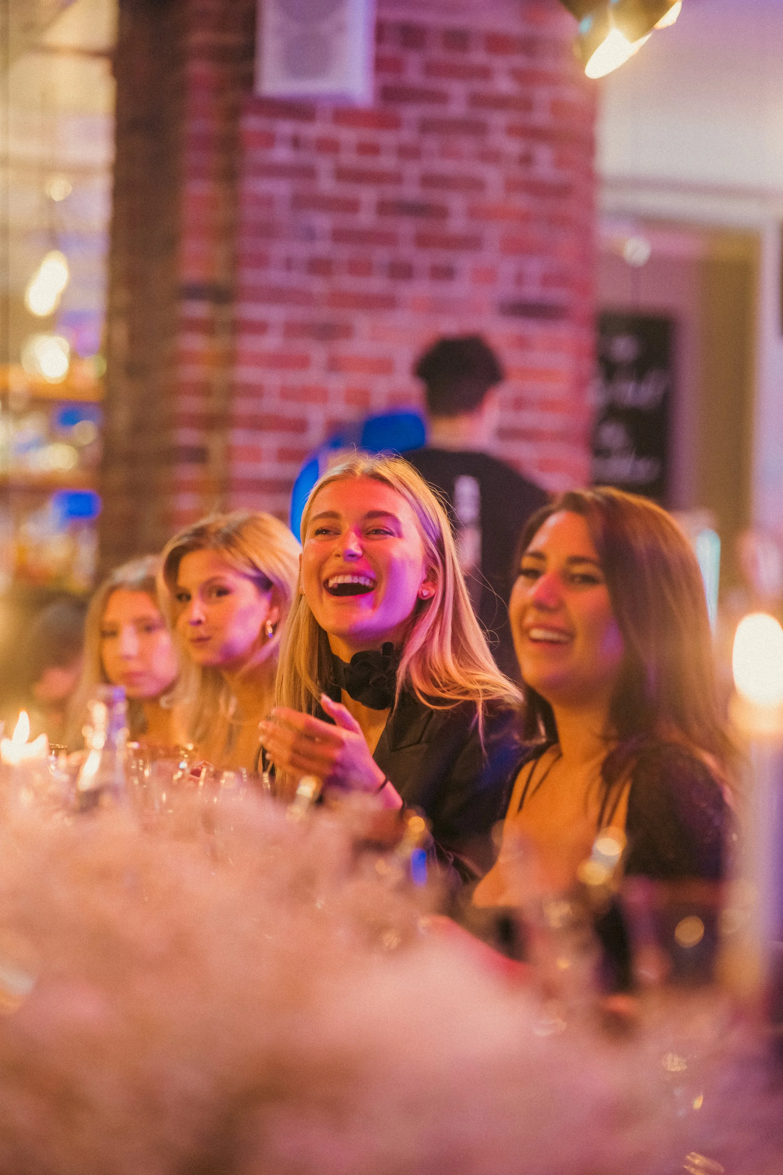 People enjoying a lively gathering or party, some smiling and engaged in conversation, with warm lighting and a brick wall in the background.