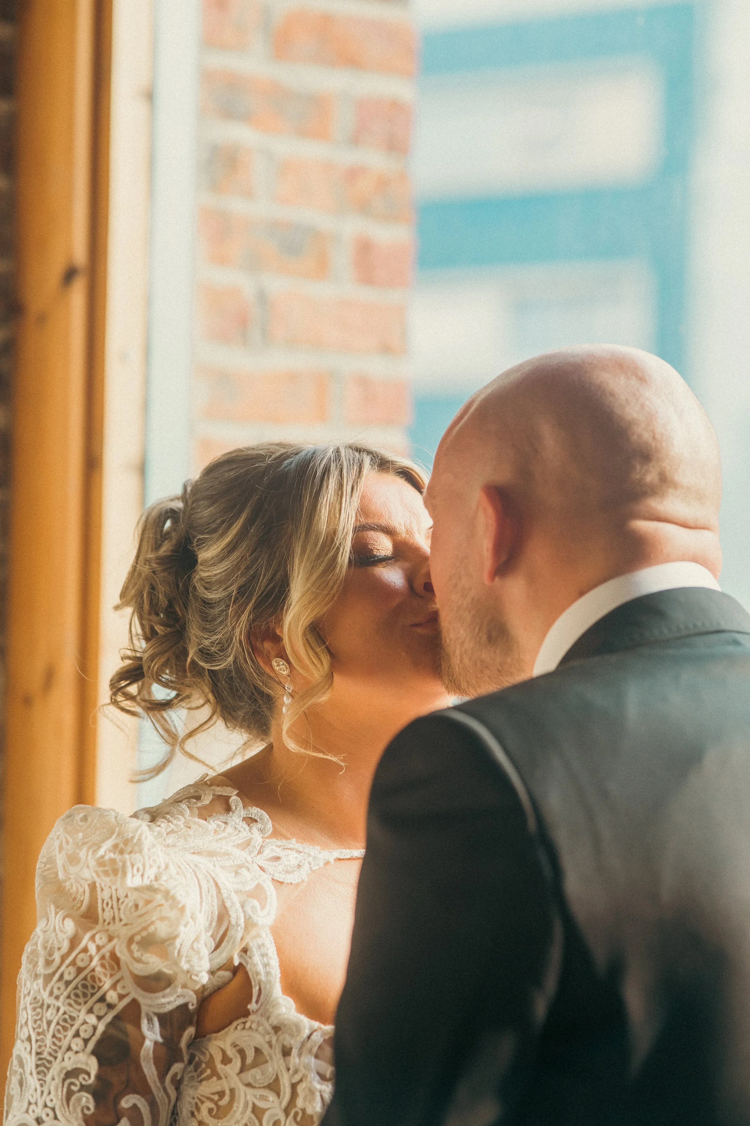 A bride and groom sharing a kiss indoors, near a window with a brick wall outside.