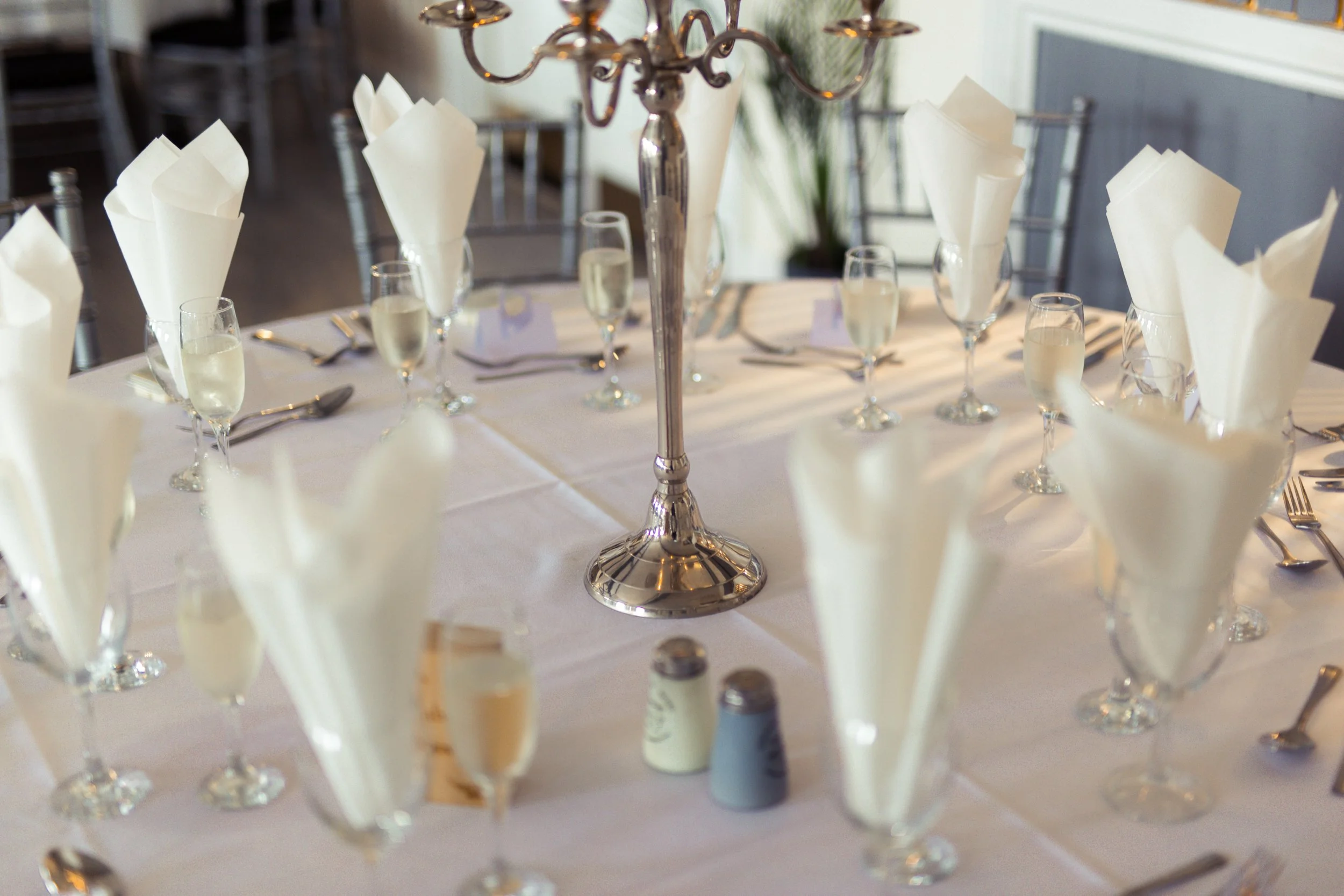 A formal dining table set with champagne glasses filled with champagne, white napkins folded in a cone shape, silverware, and a silver candle holder on a white tablecloth.