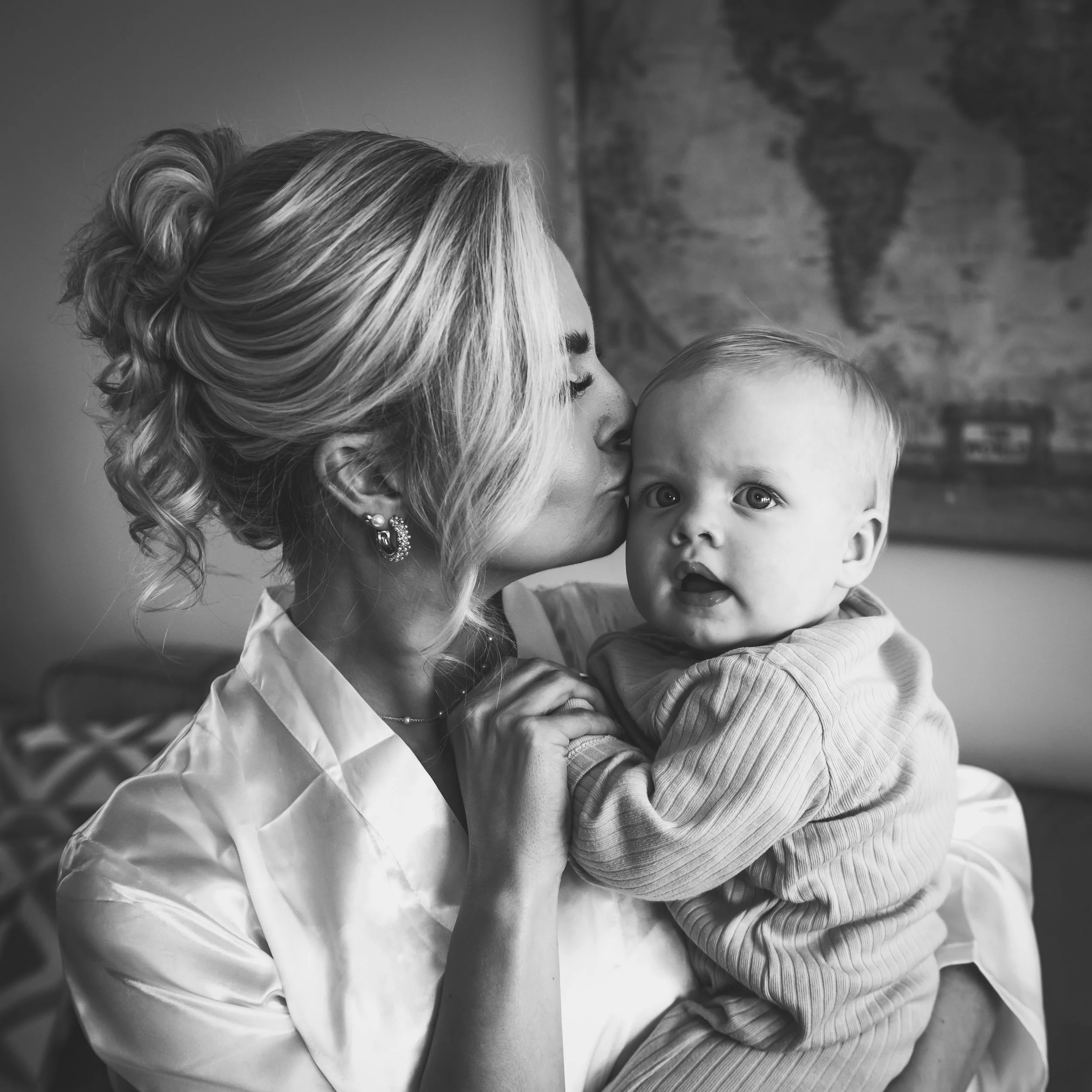 A woman with styled blonde hair and earrings is holding a baby, giving the baby a kiss on the cheek in a room with a world map on the wall.