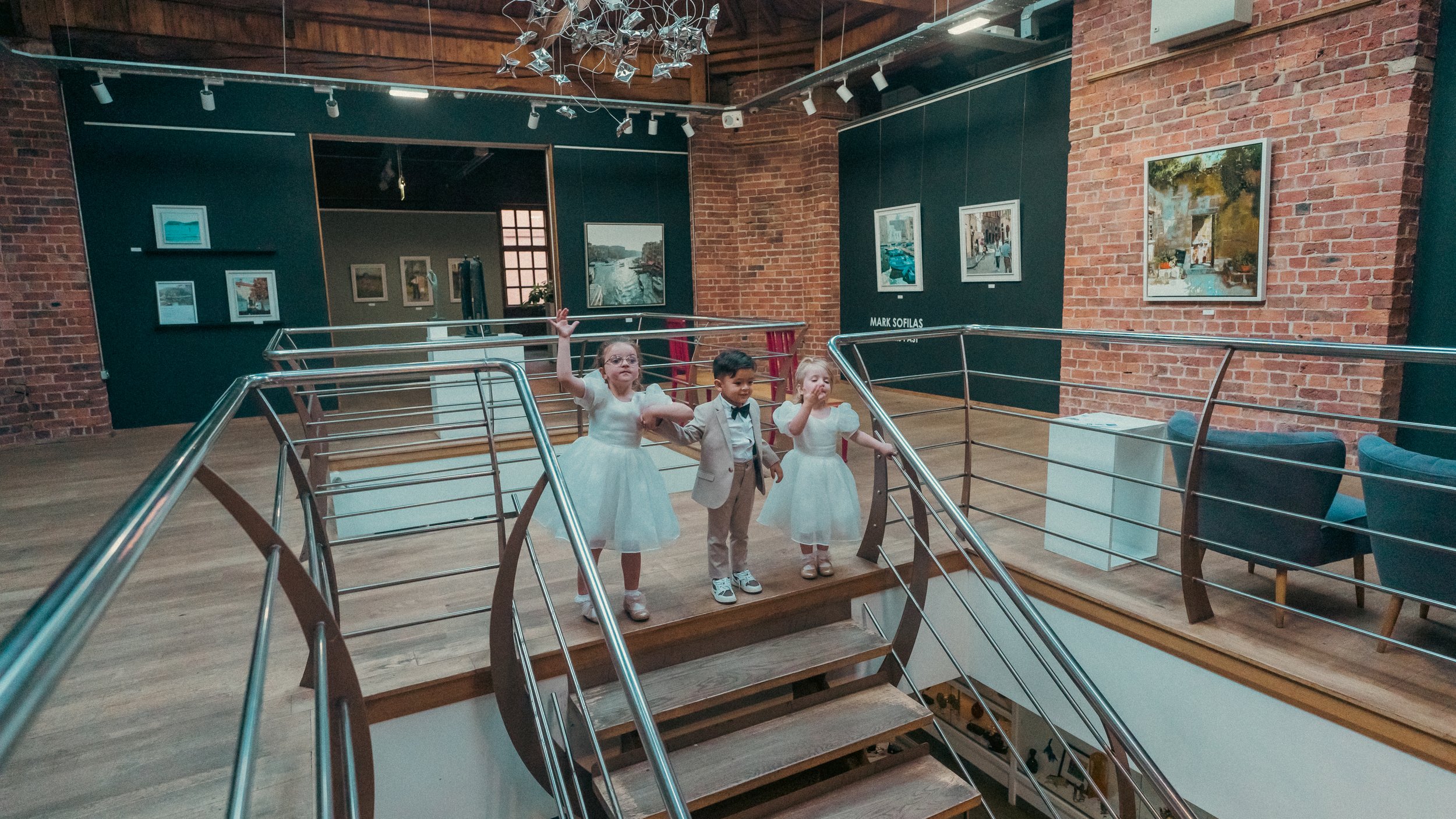 Three young children, two girls in white dresses and a boy in a suit, stand on a platform in an art gallery. Two of the girls are holding hands and the boy is in the middle. They are inside a brick-walled gallery with various paintings hanging on the