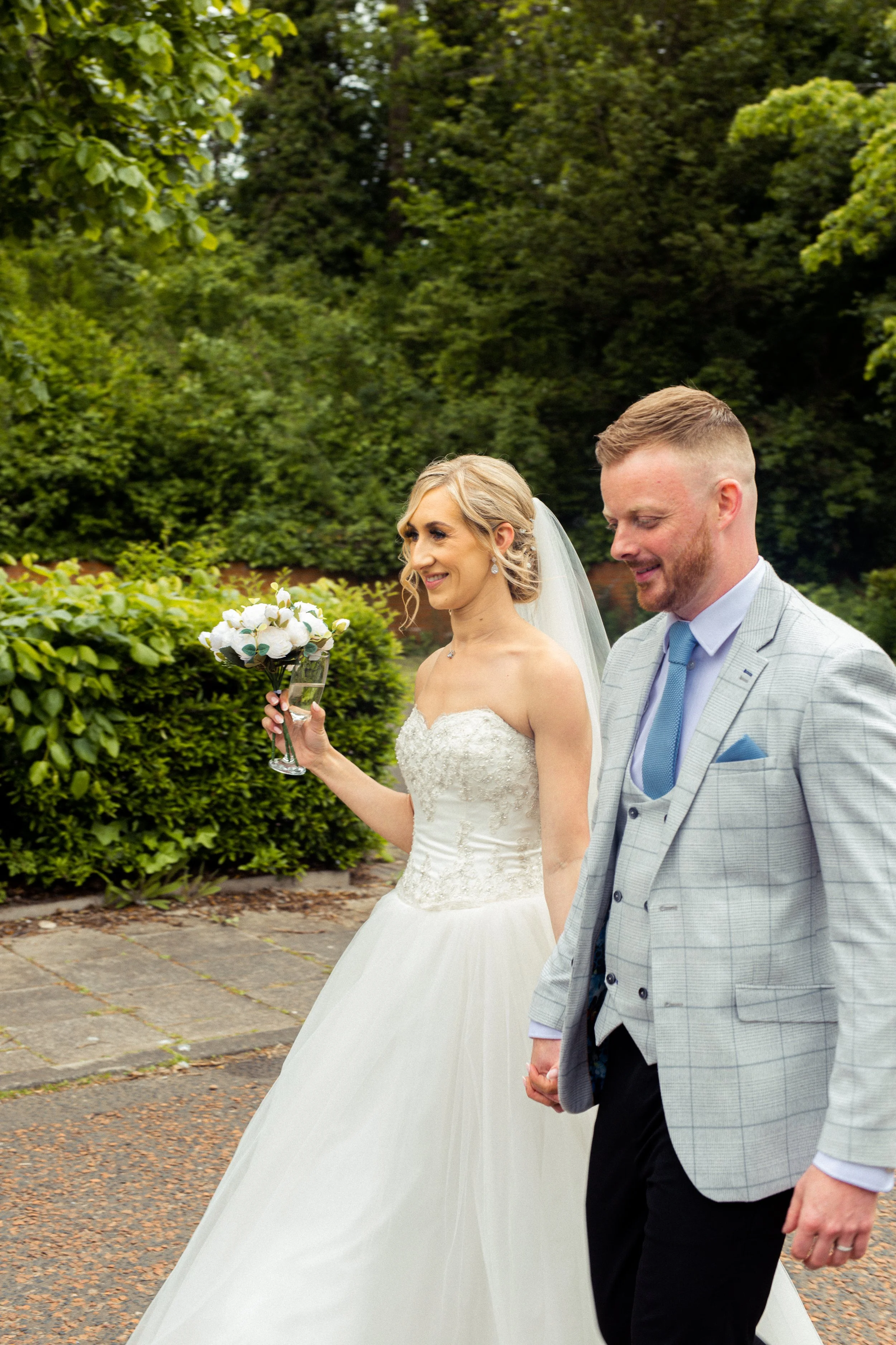 A bride and groom walking outdoors on their wedding day, with the bride holding a bouquet, surrounded by greenery.