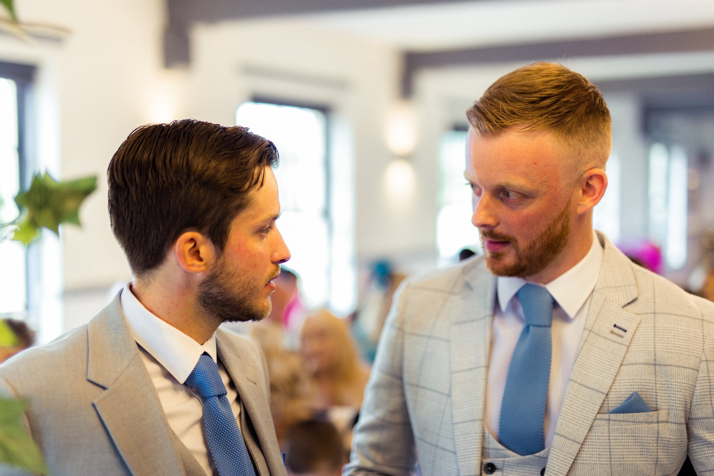 Two men in suits having a conversation at a formal event, with blurred people and windows in the background.