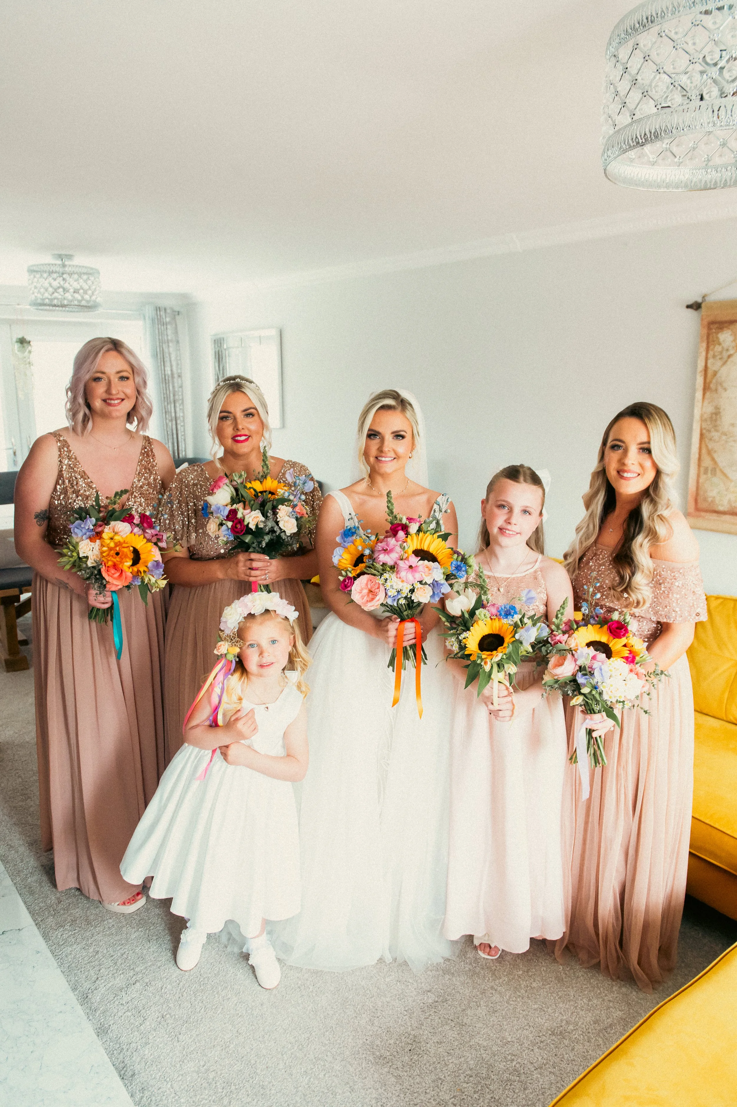 Six women and a young girl in a living room, dressed in formal attire, holding bouquets of flowers, possibly at a wedding or celebration.