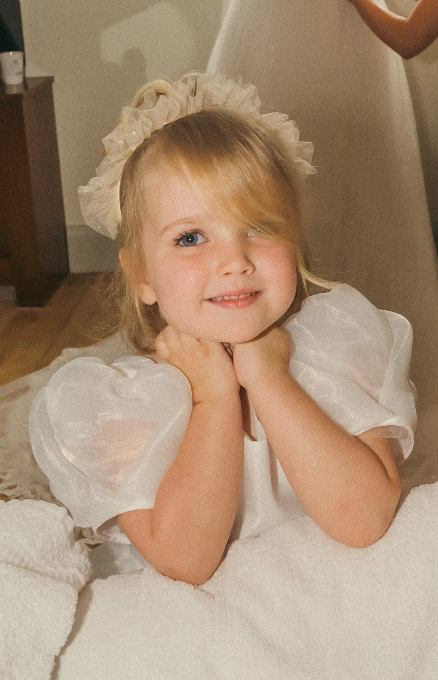 A young girl with blonde hair, blue eyes, and a bright smile, wearing a white dress with puffy sleeves and a ruffled headband, resting her chin on her hands and posing for the camera.