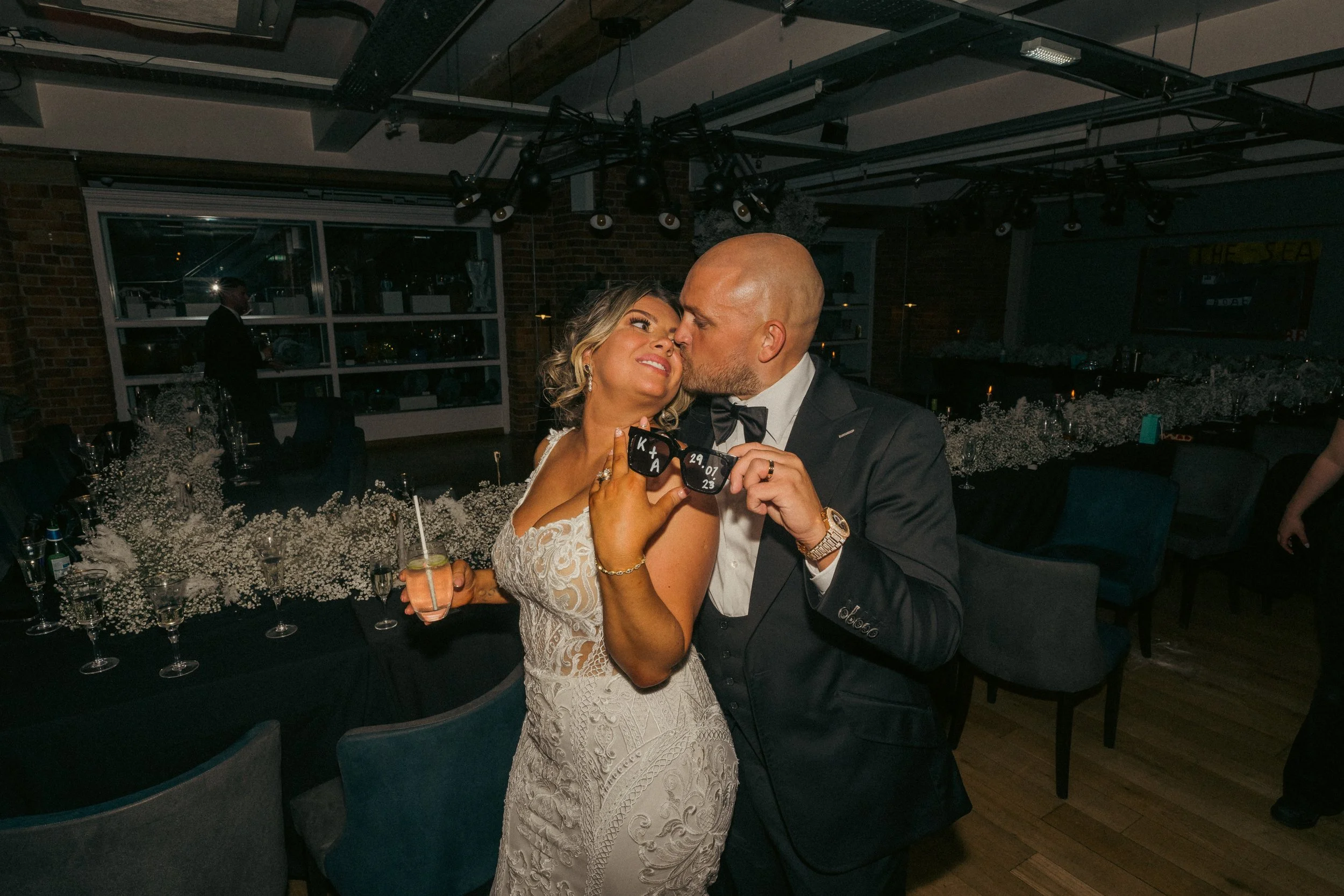 A couple dressed in wedding attire sharing a romantic moment at a wedding reception. The woman is in a white lace wedding dress, and the man is in a black tuxedo. They are holding a photo prop with their initials, wedding date, and the word 'LOVE'. T