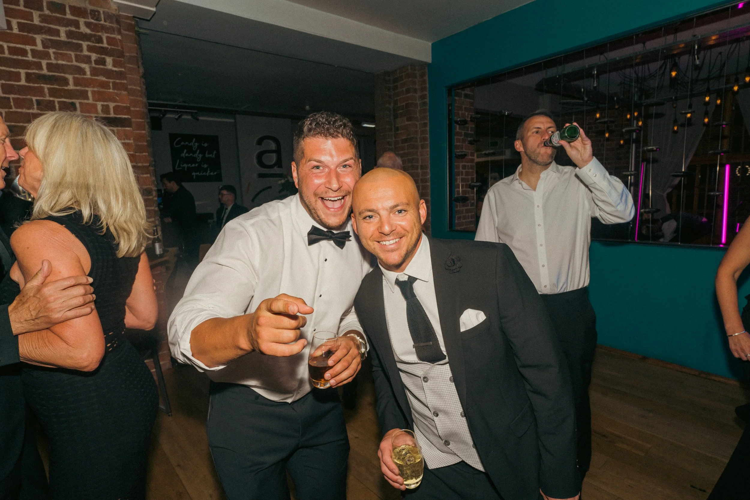 Two men in tuxedos smiling and posing for a photo at a party, one holding a glass of whiskey and the other holding a glass of a clear drink, with other guests in the background.