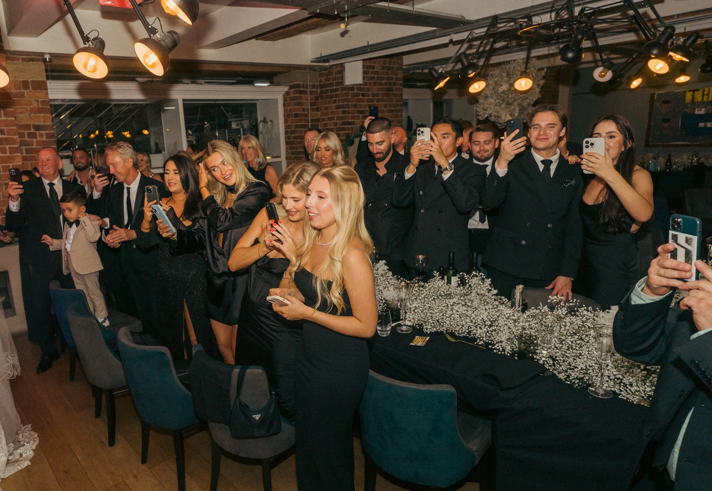 People gathered at a formal event, many are dressed in black and taking photos or videos with their smartphones. Some are smiling and looking towards the front, around a table decorated with white baby's breath flowers.