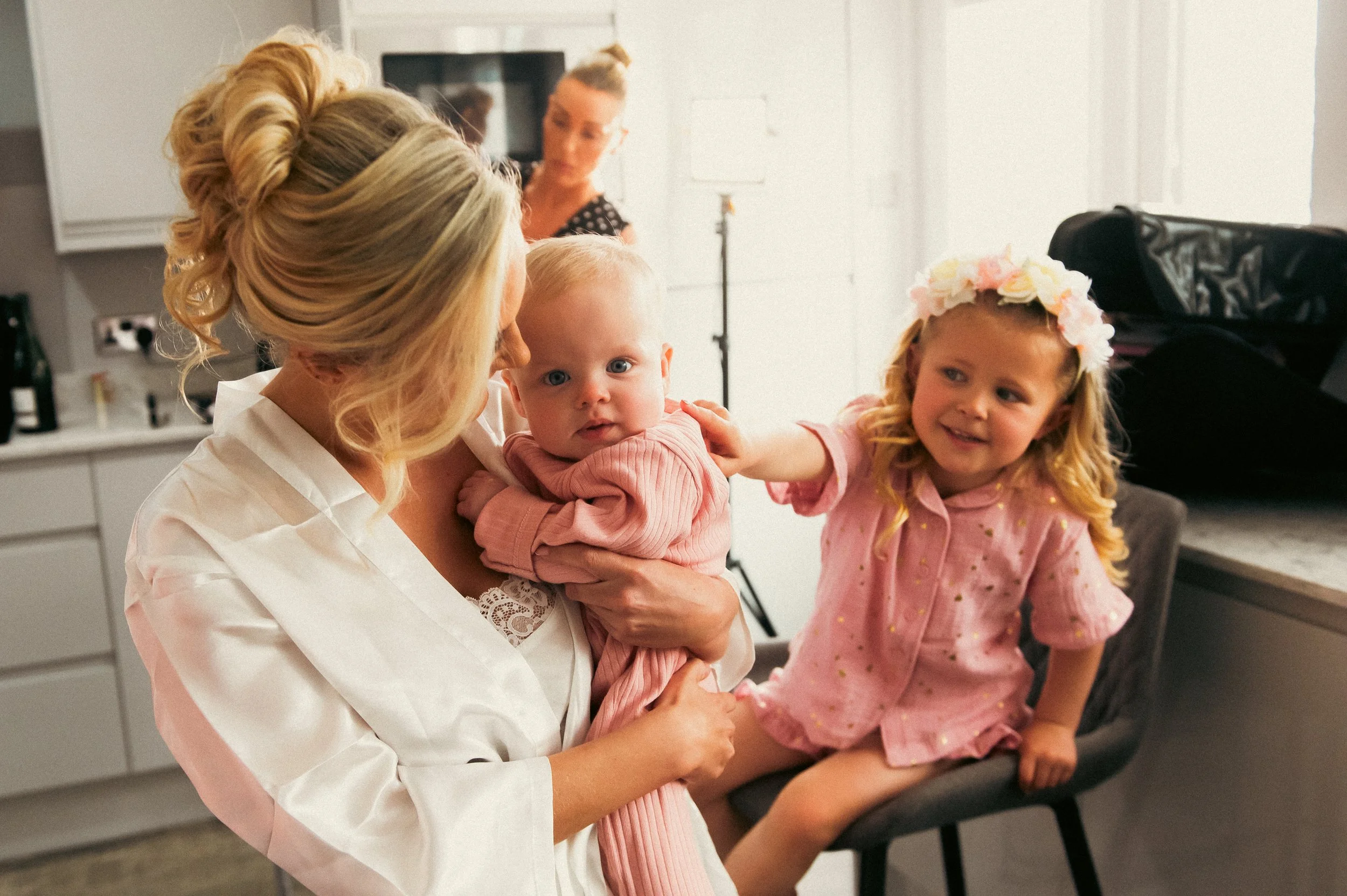 A woman with blond hair styled in an updo holding a baby girl and a young girl with blond hair and a floral headband, in a room with white cabinets and a TV in the background.
