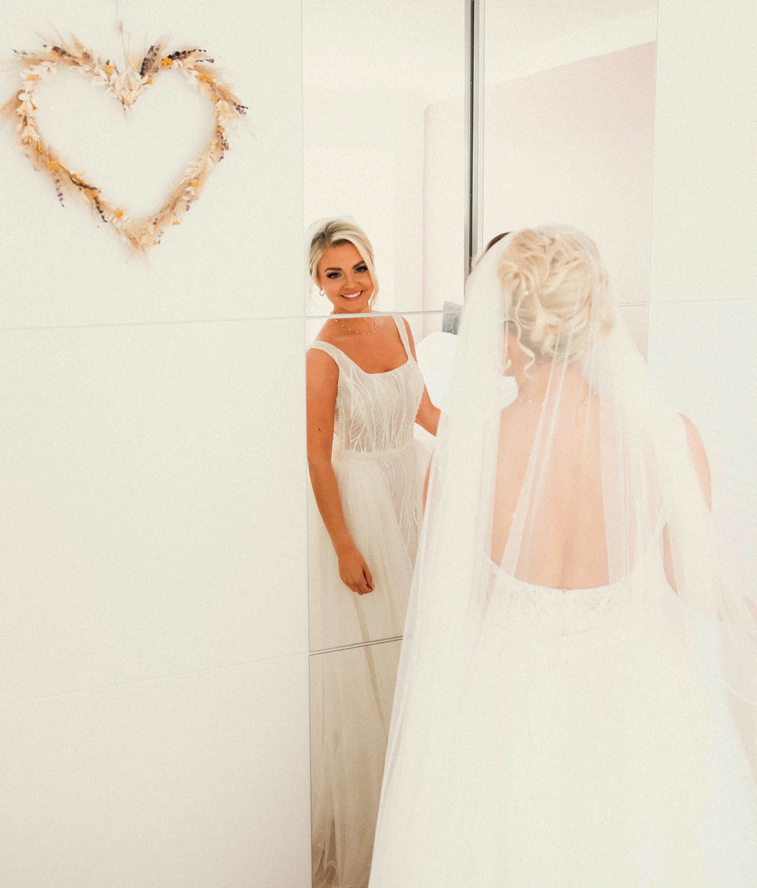 Bride in a wedding dress looking at herself in a mirror, with a heart-shaped flower arrangement on the wall