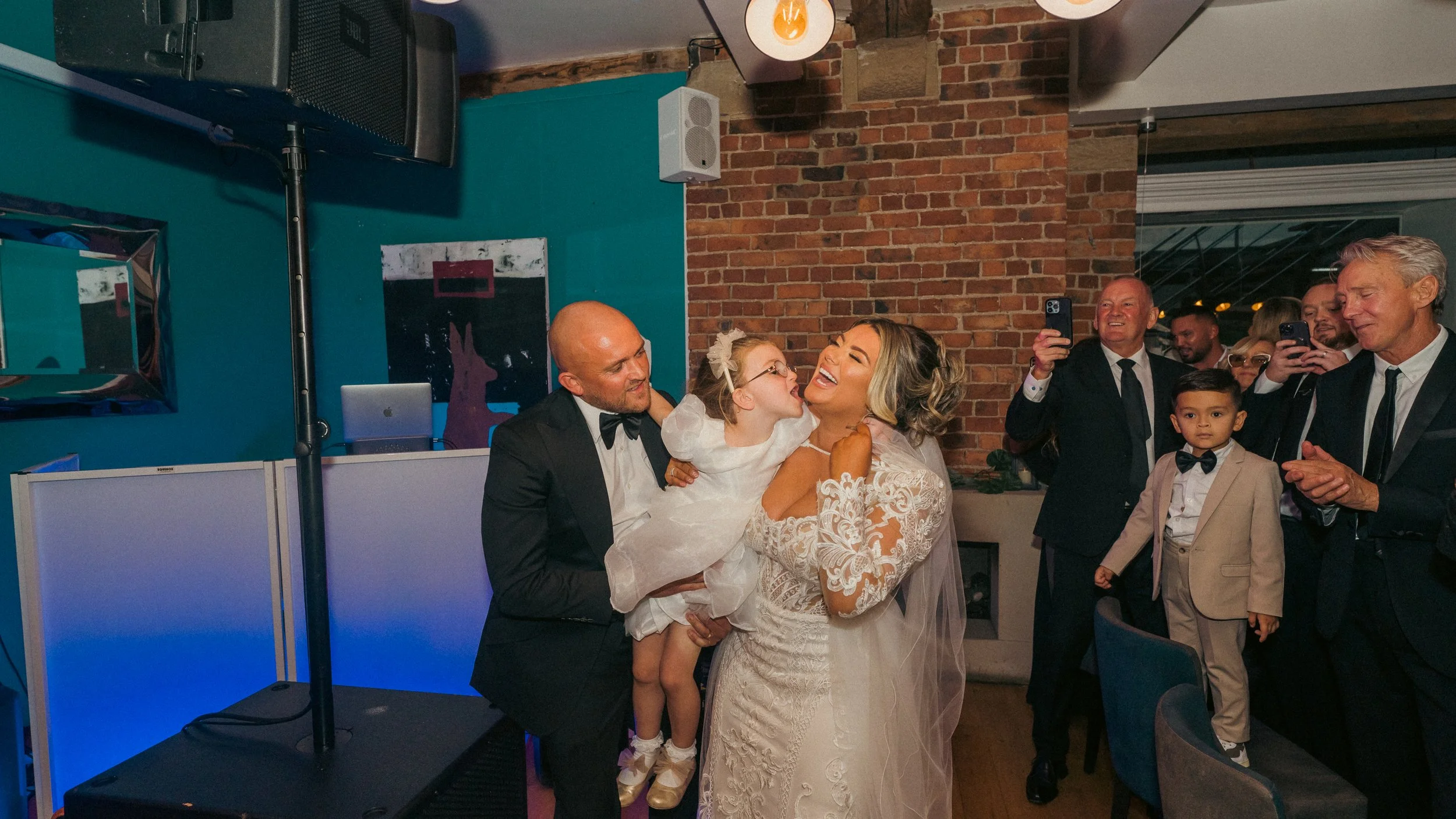 A group of people at a wedding reception, including a bride in a lace wedding dress, a groom in a tuxedo, and a young girl in a white dress, sharing a joyful moment with others taking photos and observing.