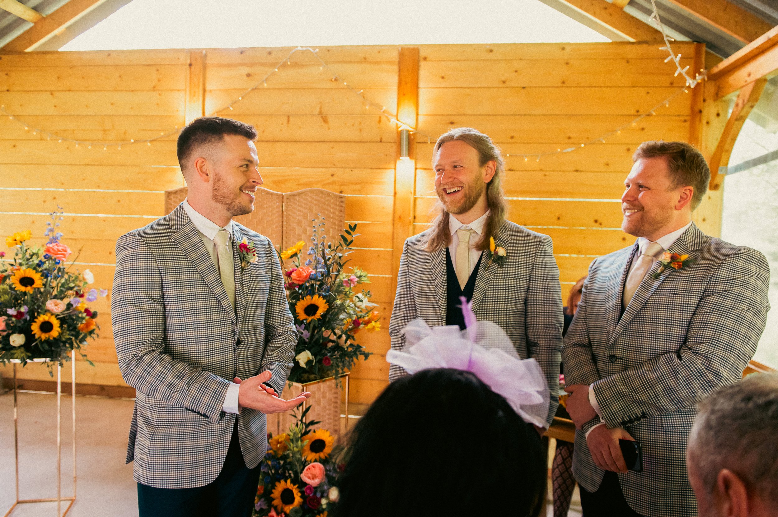 Three men in checkered suits stand inside a wooden wedding venue, smiling at each other. Floral arrangements and string lights decorate the background.