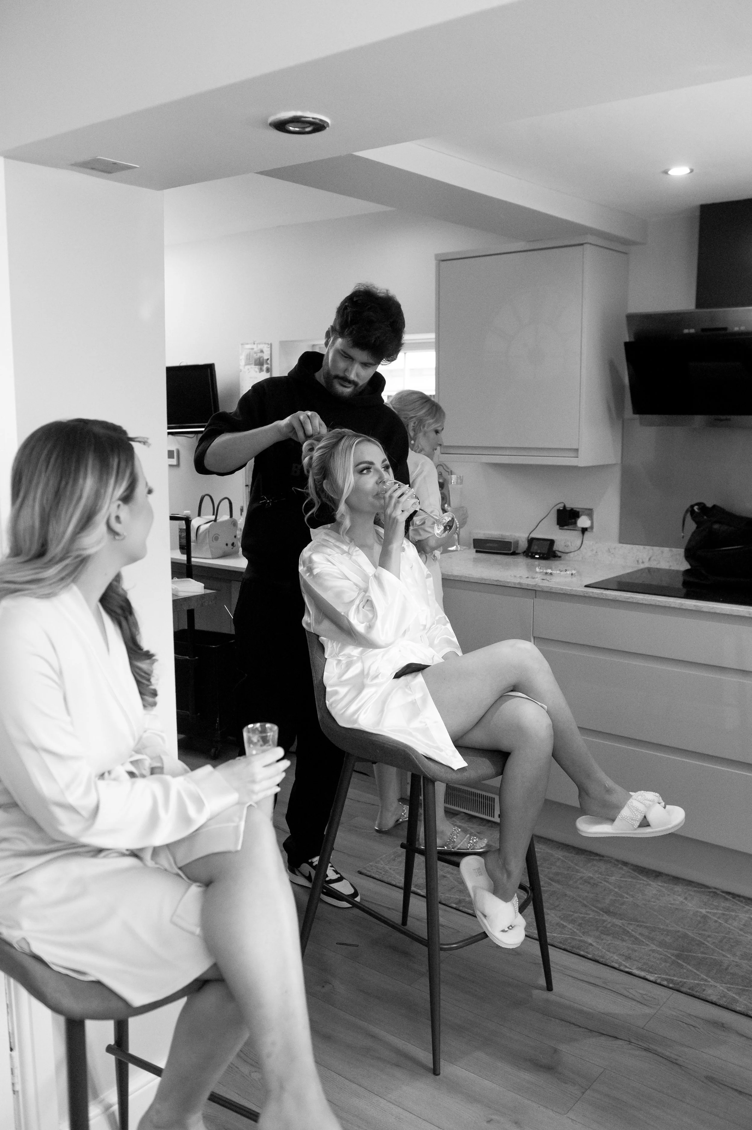 Women getting ready, with one woman sitting on a stool having her hair styled while wearing a satin robe, another woman sitting nearby holding a drink, and a woman in the background drinking from a glass in a kitchen.