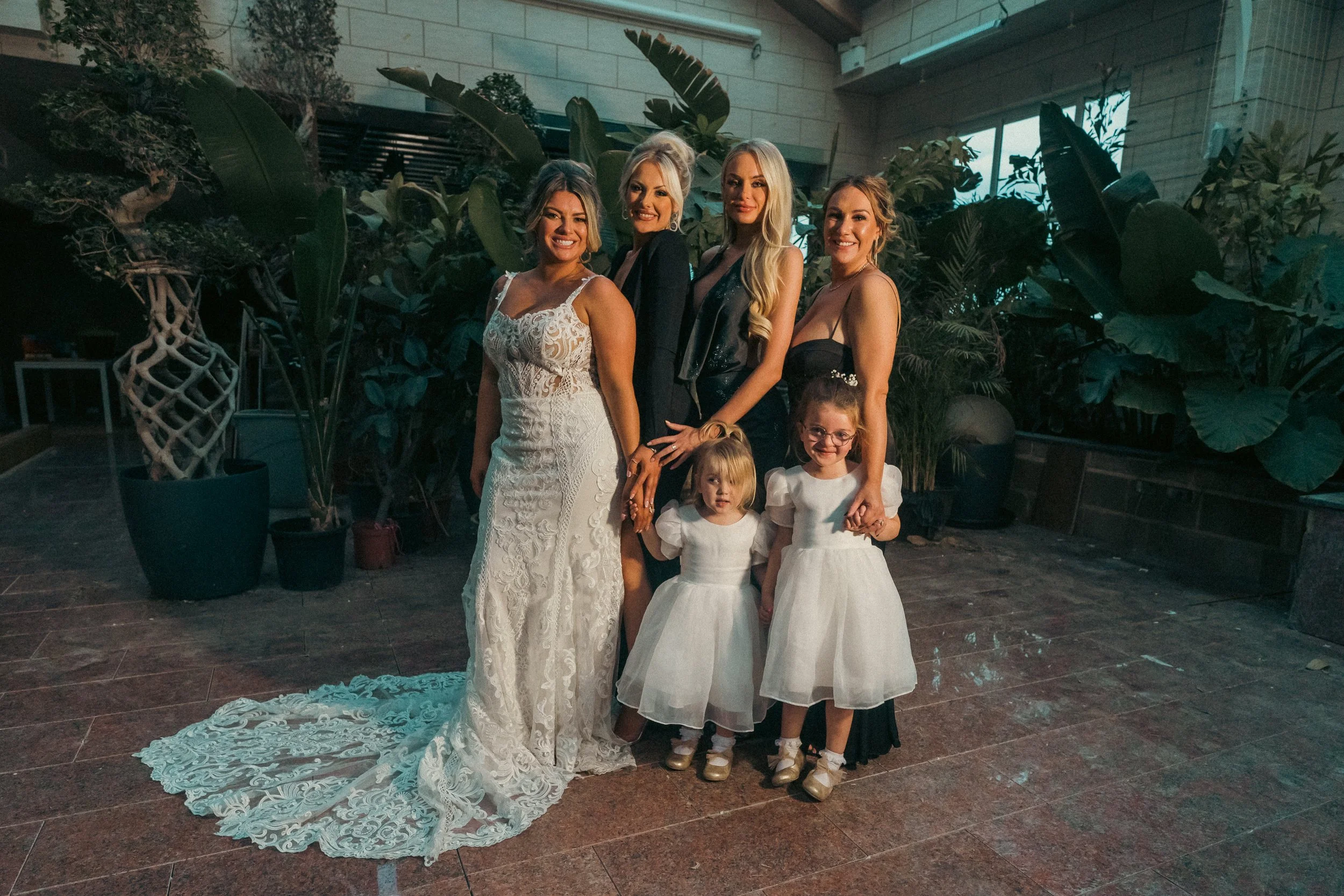 Group of five women and two young girls in formal attire standing in front of green plants indoors.