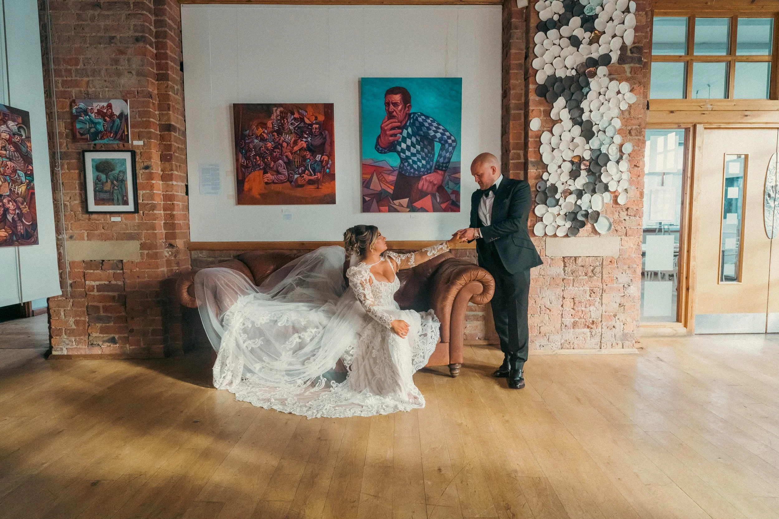 A bride in a white lace wedding dress and veil is sitting on a vintage brown leather couch, reaching out her hand to a man in a black tuxedo, who is standing and holding her hand. They are in an art gallery with brick walls, colorful paintings, and m