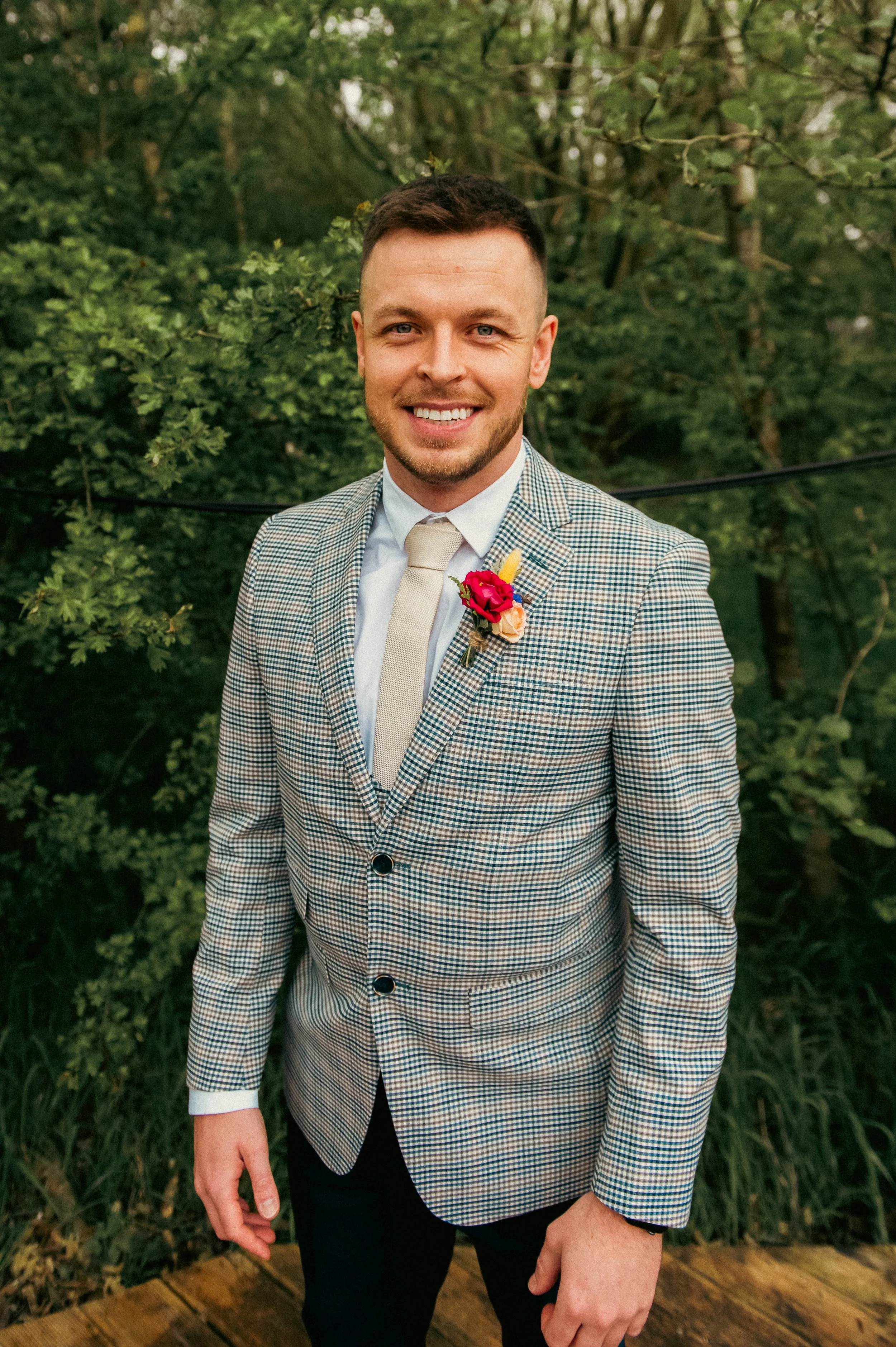 A smiling man wearing a checked blazer, white shirt, and a beige tie with a pink and red boutonniere, outdoors with green trees in the background.