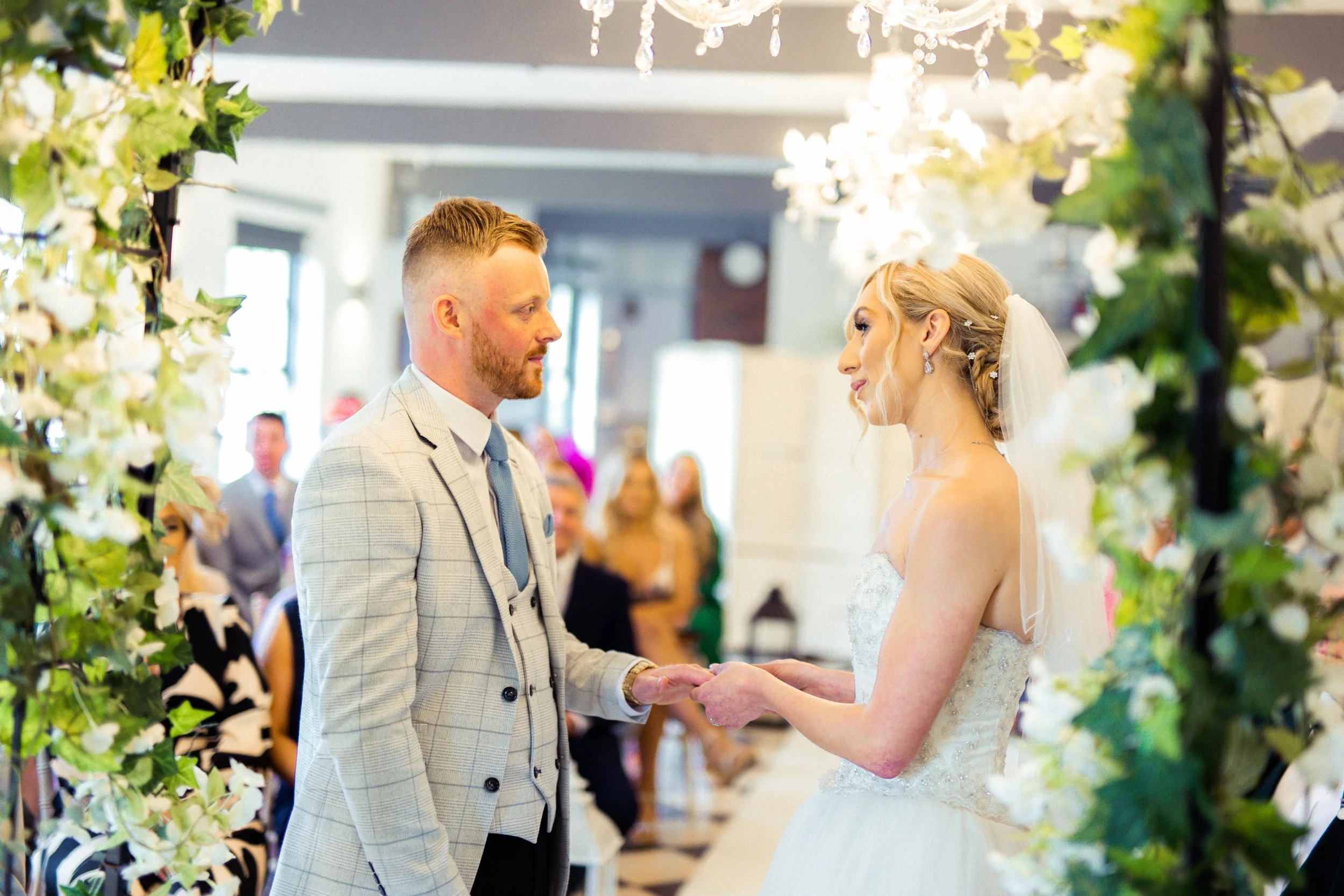 A bride and groom standing face to face during their wedding ceremony, holding hands, surrounded by floral decorations and guests in the background.