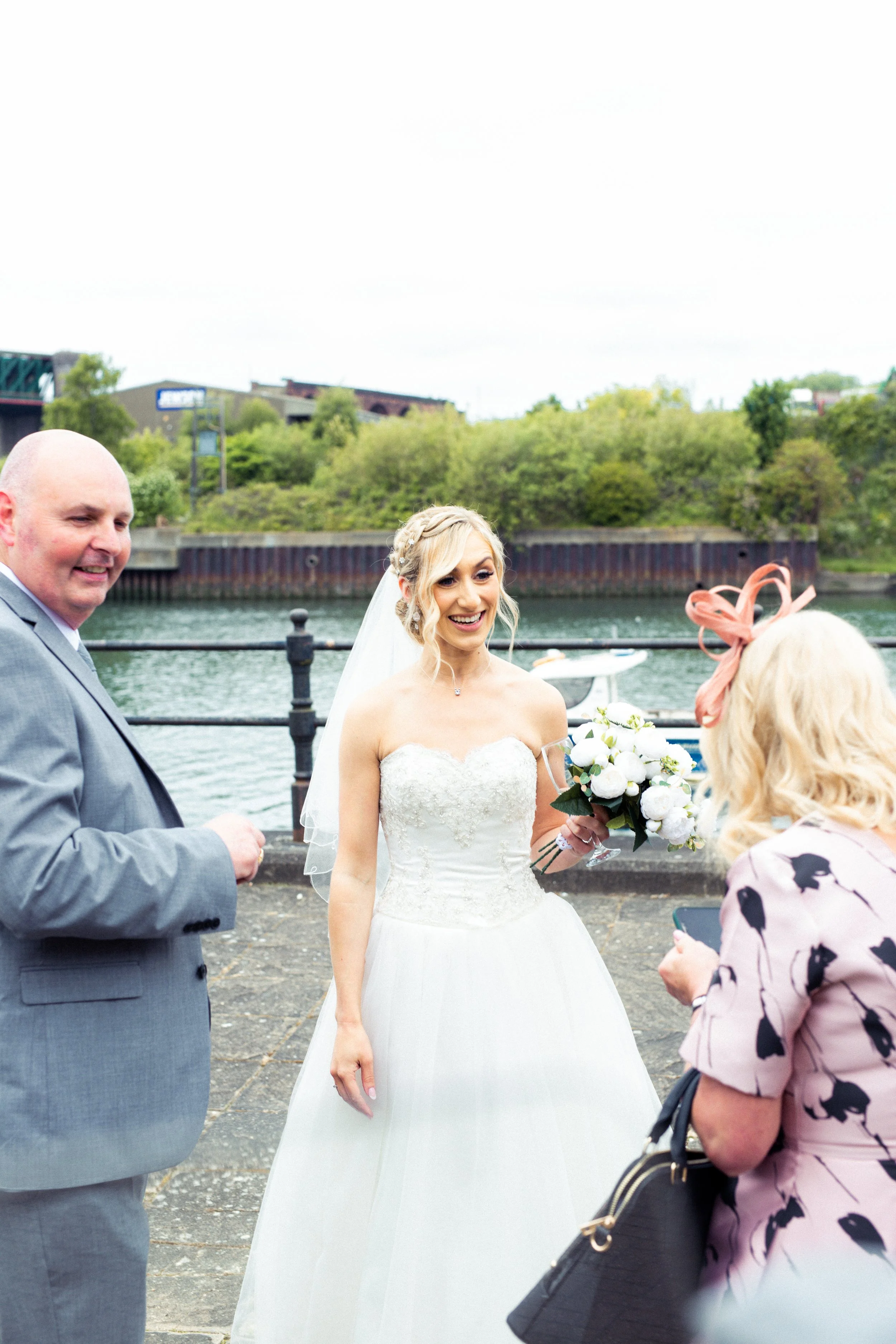 Bride in a white wedding dress holding a bouquet of white flowers, smiling and talking to two guests near a waterfront with boats and greenery in the background.